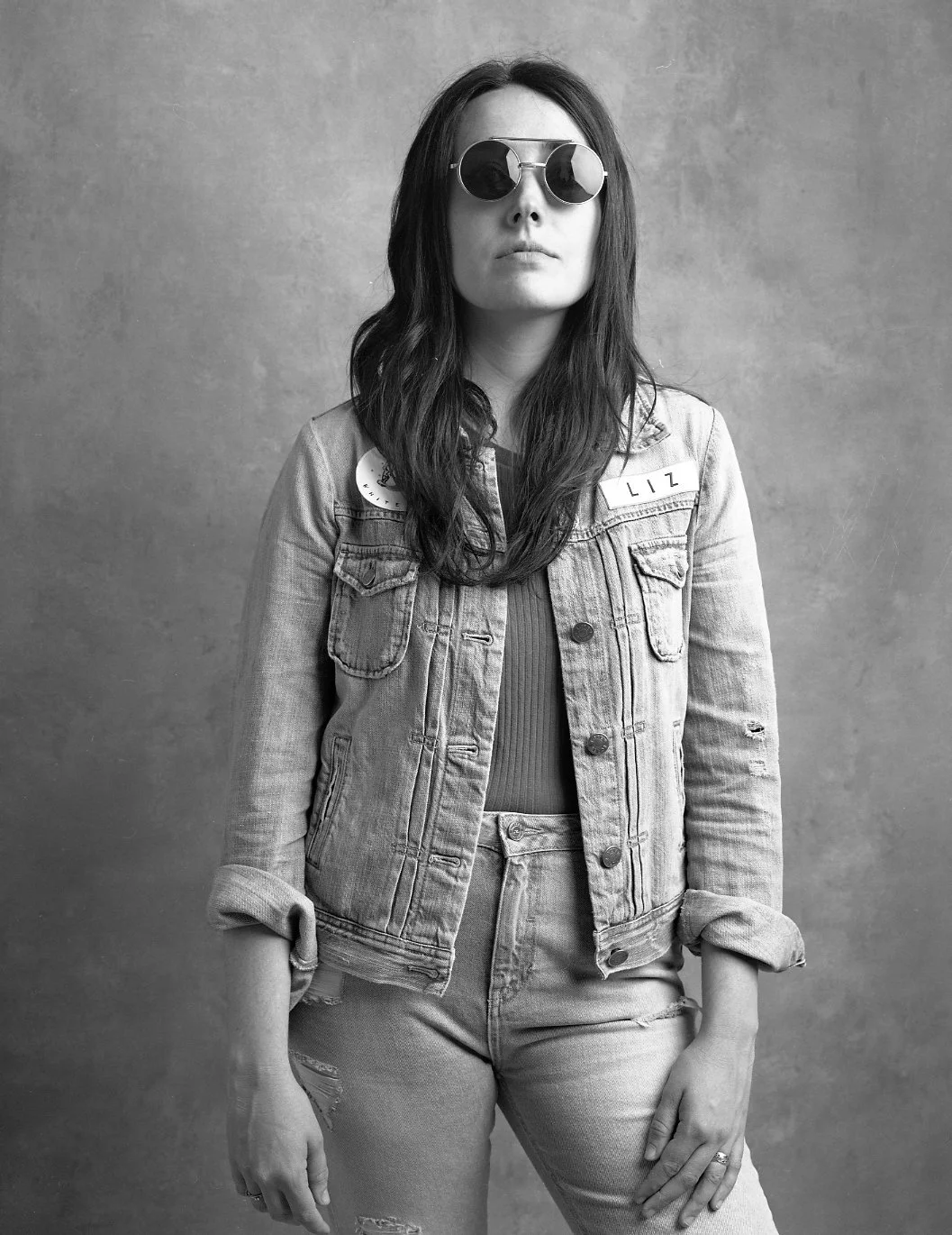 Black and white photo of a woman with long hair wearing sunglasses, a denim jacket with patches, a ribbed top, and jeans, standing against a plain background.