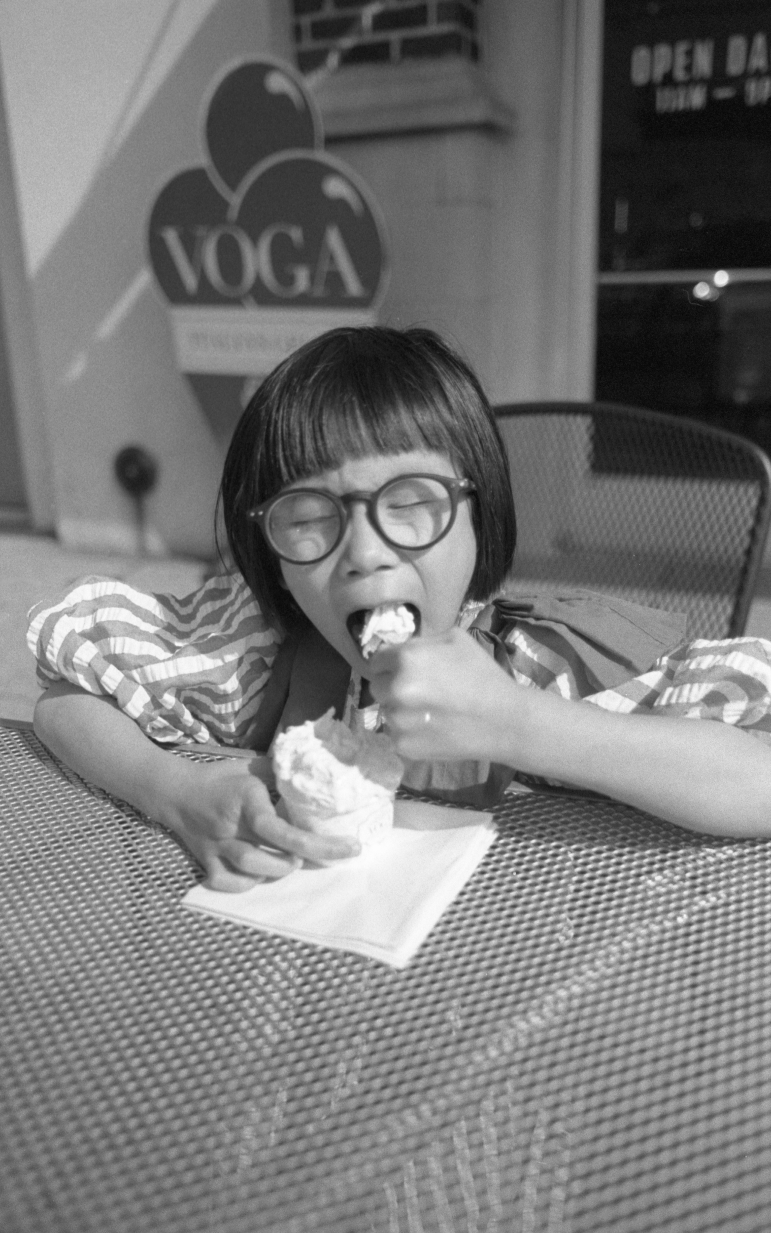 A young girl with short hair, glasses, and a striped shirt is sitting at a table, eating ice cream with her eyes closed. There is a sign with the word 'YOGA' and part of a window or door in the background.