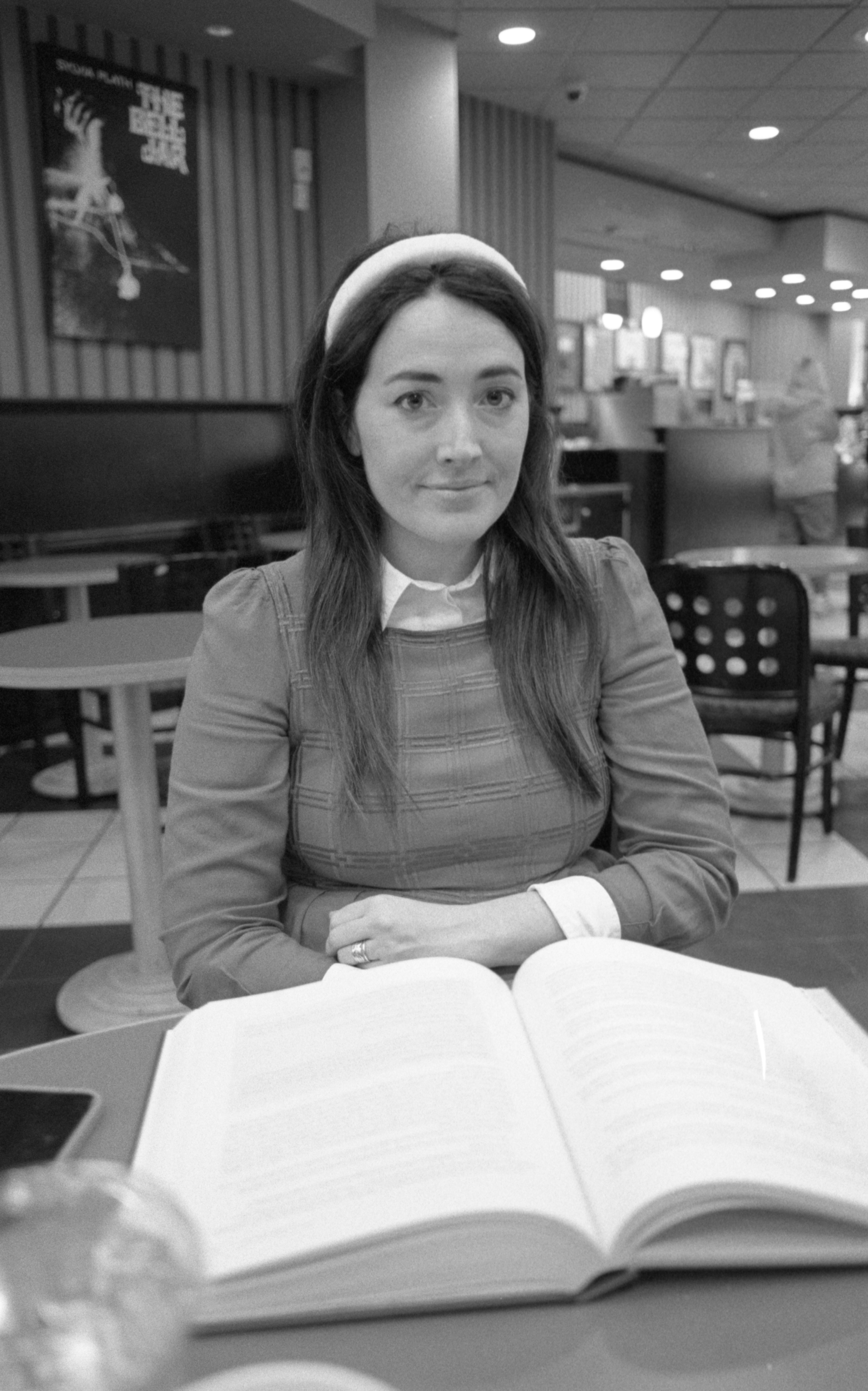 A woman sitting at a table in a restaurant or cafe, facing the camera with an open book in front of her, wearing a headband and a checked dress, in a black-and-white photo.