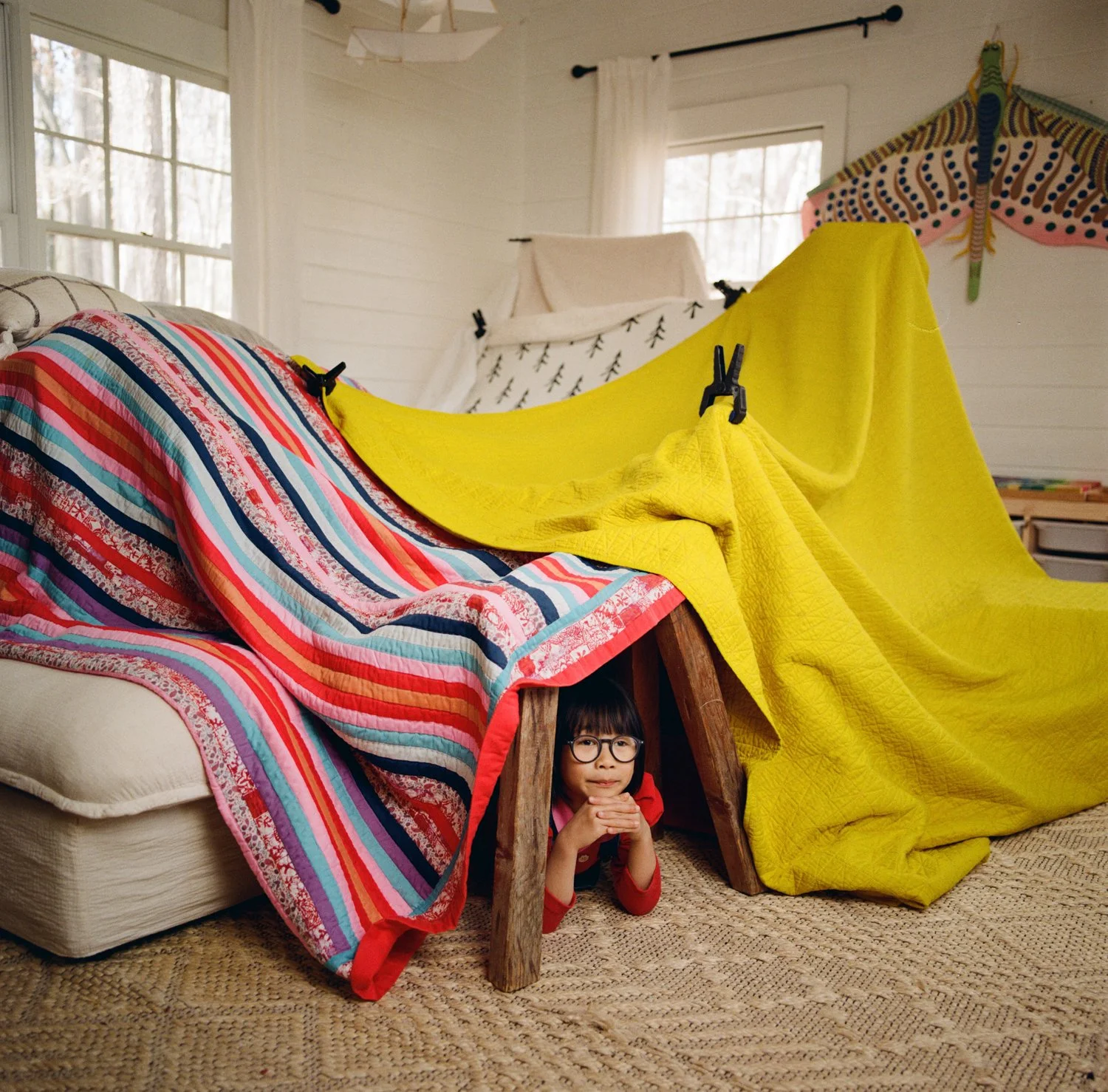 A young girl with glasses peeks out from under a sofa with a colorful striped quilt draped over it in a cozy, well-lit living room.