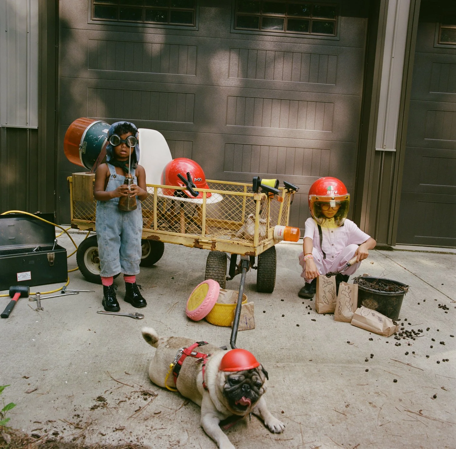 Two children wearing helmets and safety gear pose with a small vehicle and tools in a driveway. One child stands holding a bottle, the other crouches near a box of rocks with a helmet and goggles. A bulldog wearing a helmet lies on the ground nearby.