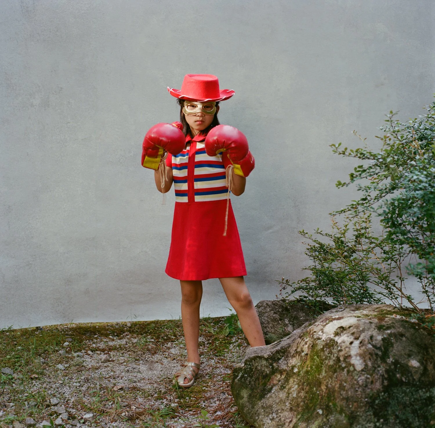 Young girl dressed as a boxer with boxing gloves, a red dress, a striped shirt, a red hat, and sunglasses standing outdoors next to a large rock and green bushes.