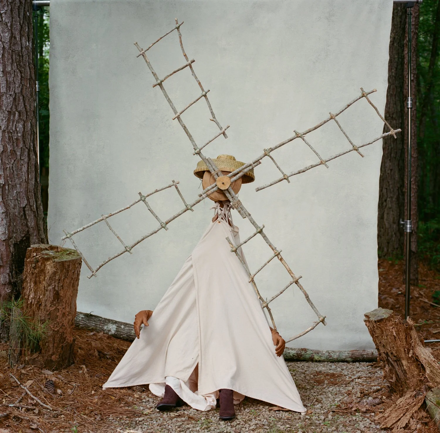 A person dressed as a scarecrow with a beige dress, wooden hands, a straw hat, and a wooden cross formed by sticks on their back, standing outdoors among trees with a plain white backdrop.