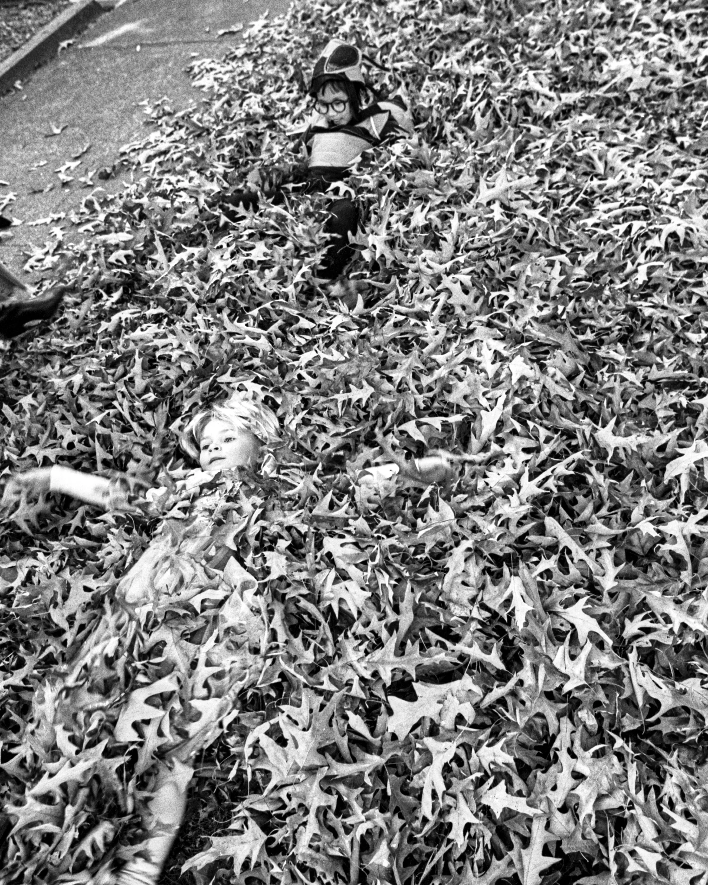 Two children lying in a large pile of fallen autumn leaves, one with glasses and the other with lighter hair, on a sidewalk.