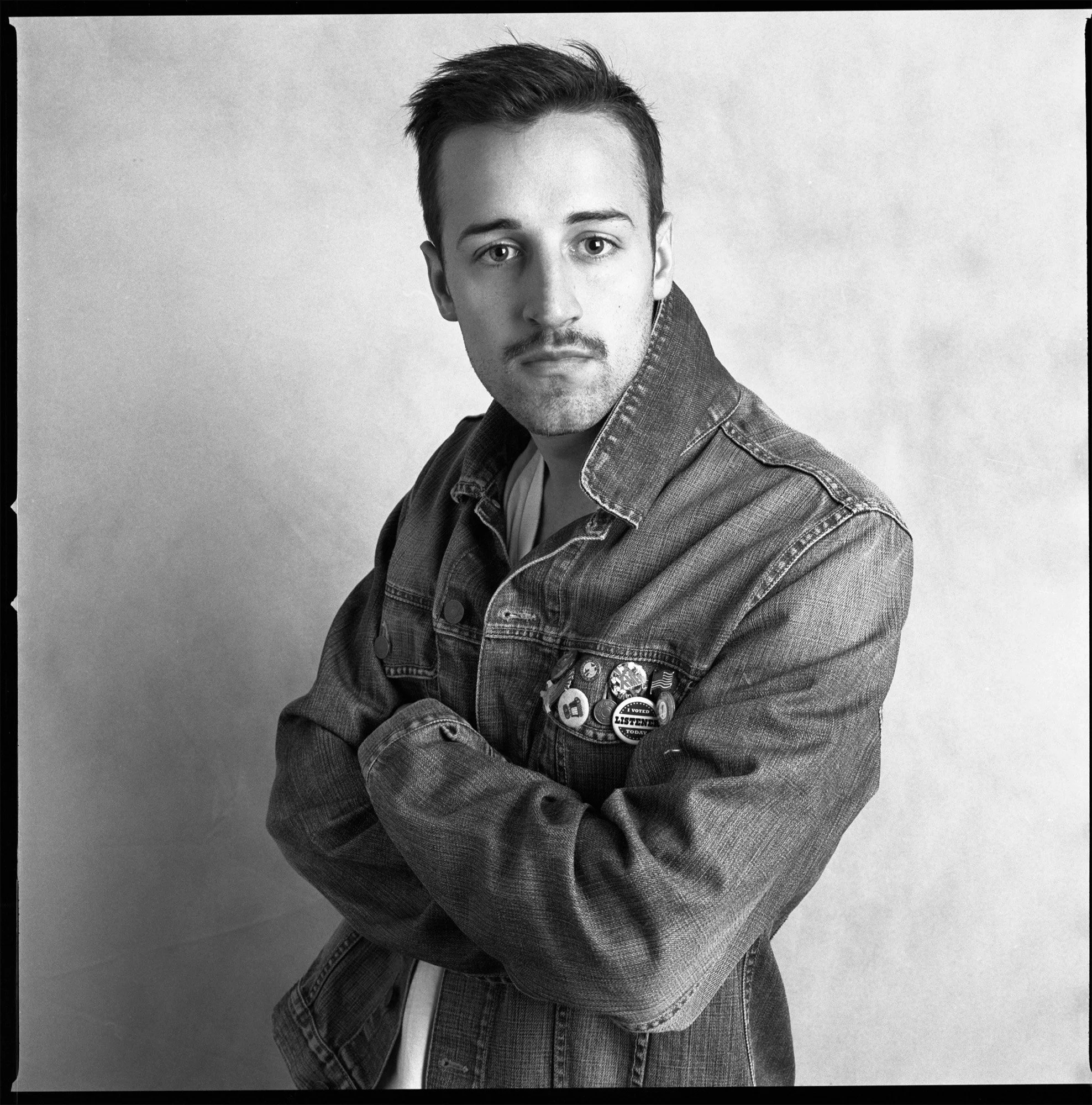 Black and white photo of a young man with short hair and a mustache, wearing a denim jacket with pins, standing with arms crossed against a plain background.