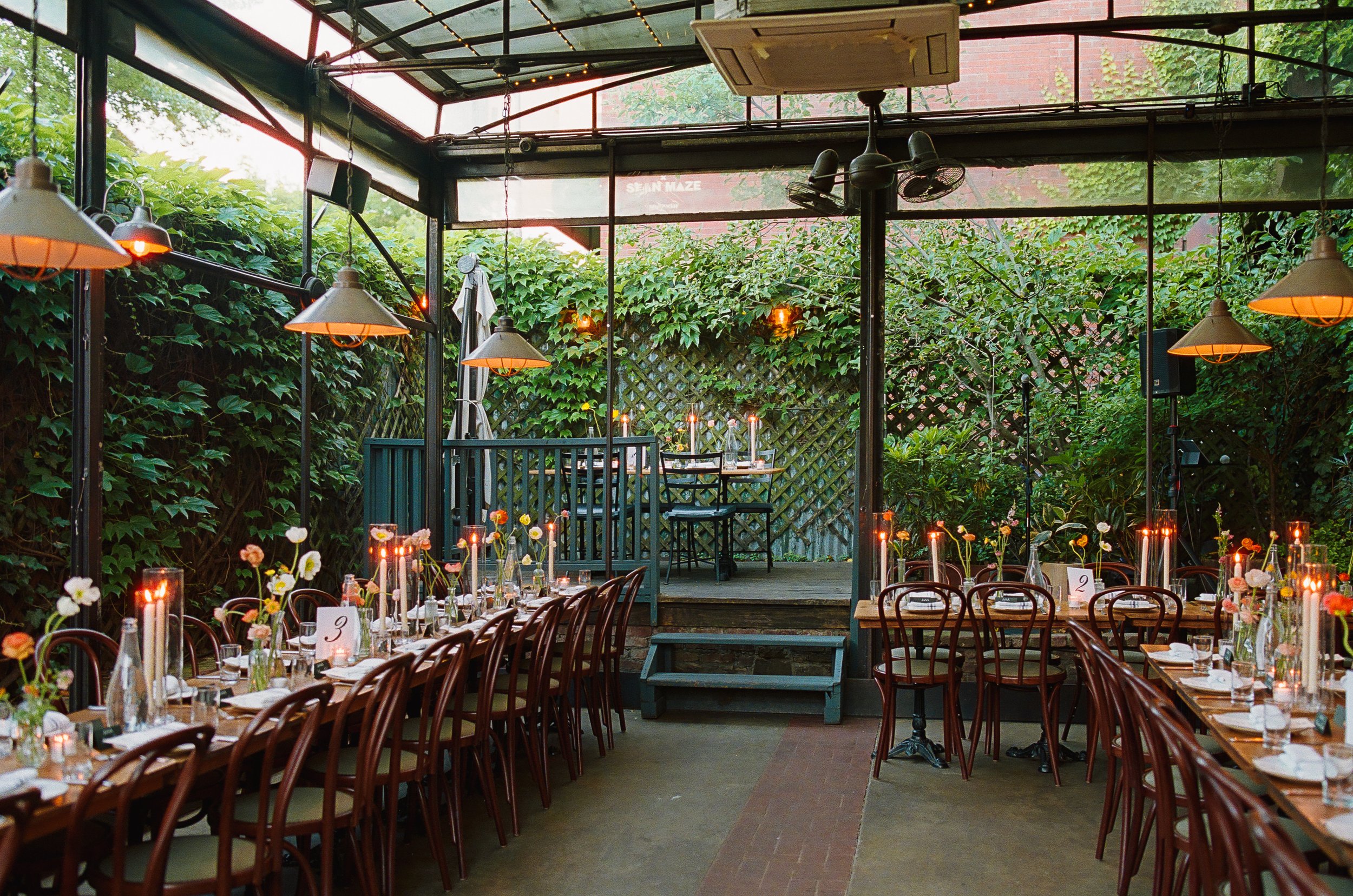 A cozy outdoor dining area decorated for an event with tables set with flowers, candles, and tableware. The area is surrounded by greenery, and there is a covered structure overhead with hanging lights and fans. Elegant chairs are arranged at the tab