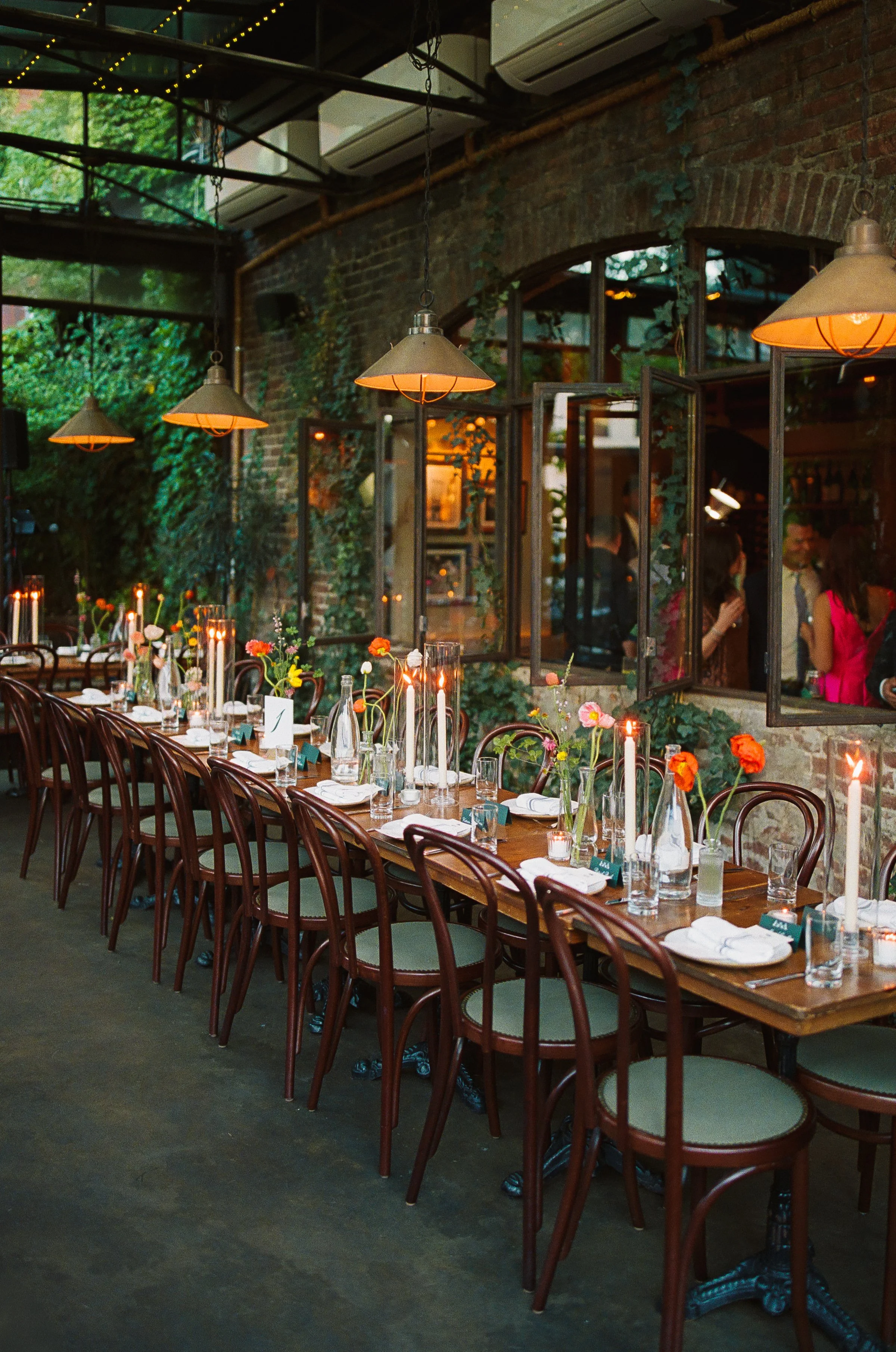 A restaurant dining area with a long wooden table set with glasses, plates, napkins, and candles. There are wooden chairs around the table. The restaurant has an industrial style with brick walls, open windows, hanging lamps, and greenery.