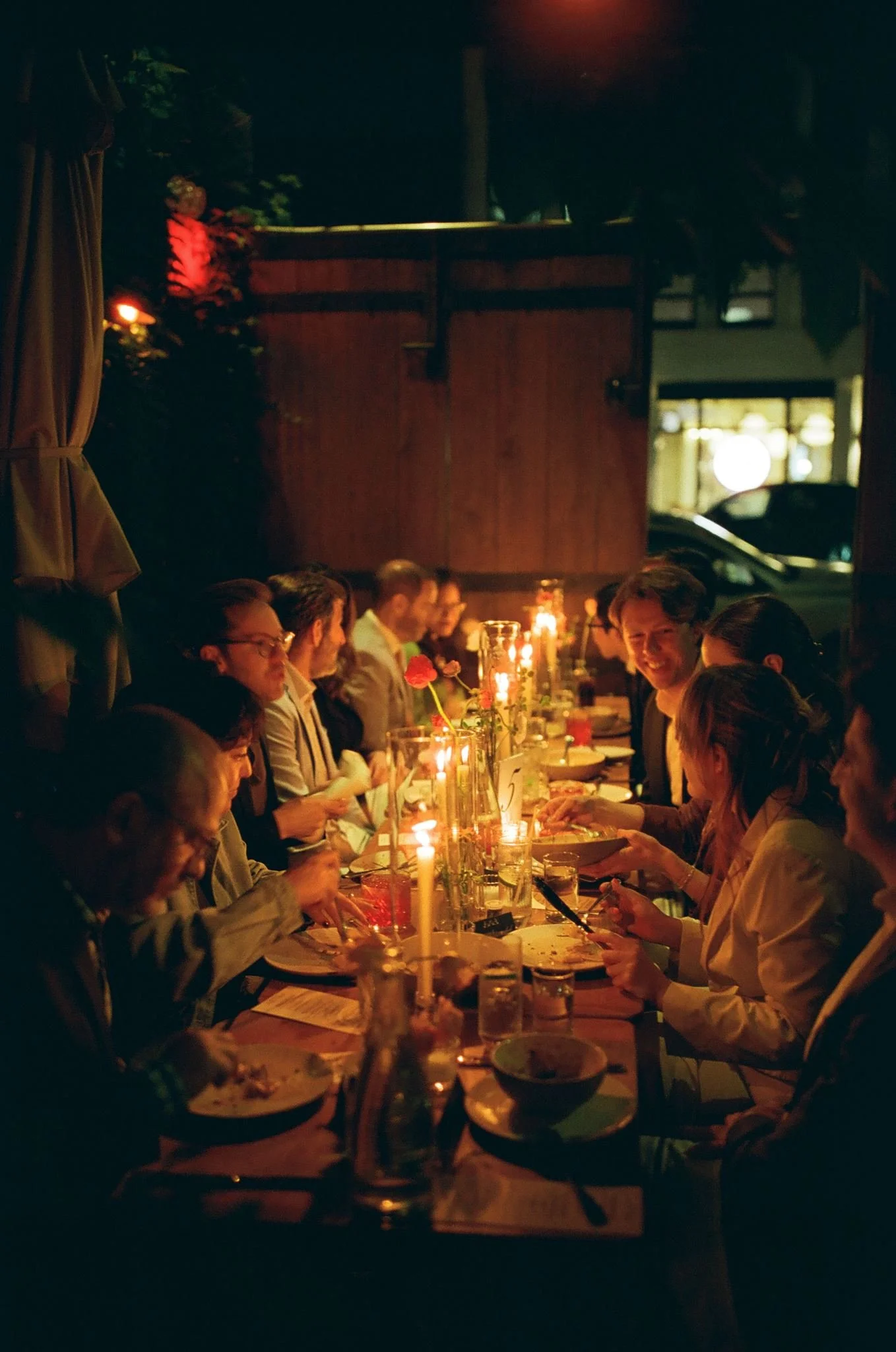 People sitting at a long dinner table in a dimly lit restaurant, illuminated by candlelight, with wearing formal attire and enjoying a meal.