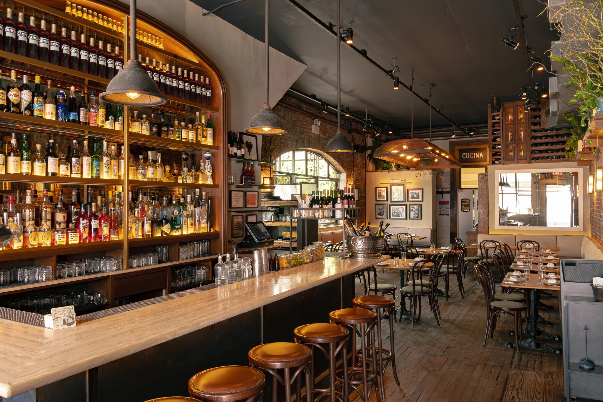 Interior of a modern restaurant or bar with a large wooden counter, shelves filled with various bottles of alcohol, and multiple tables and chairs set for dining. Industrial-style lighting and brick walls add to the ambiance.