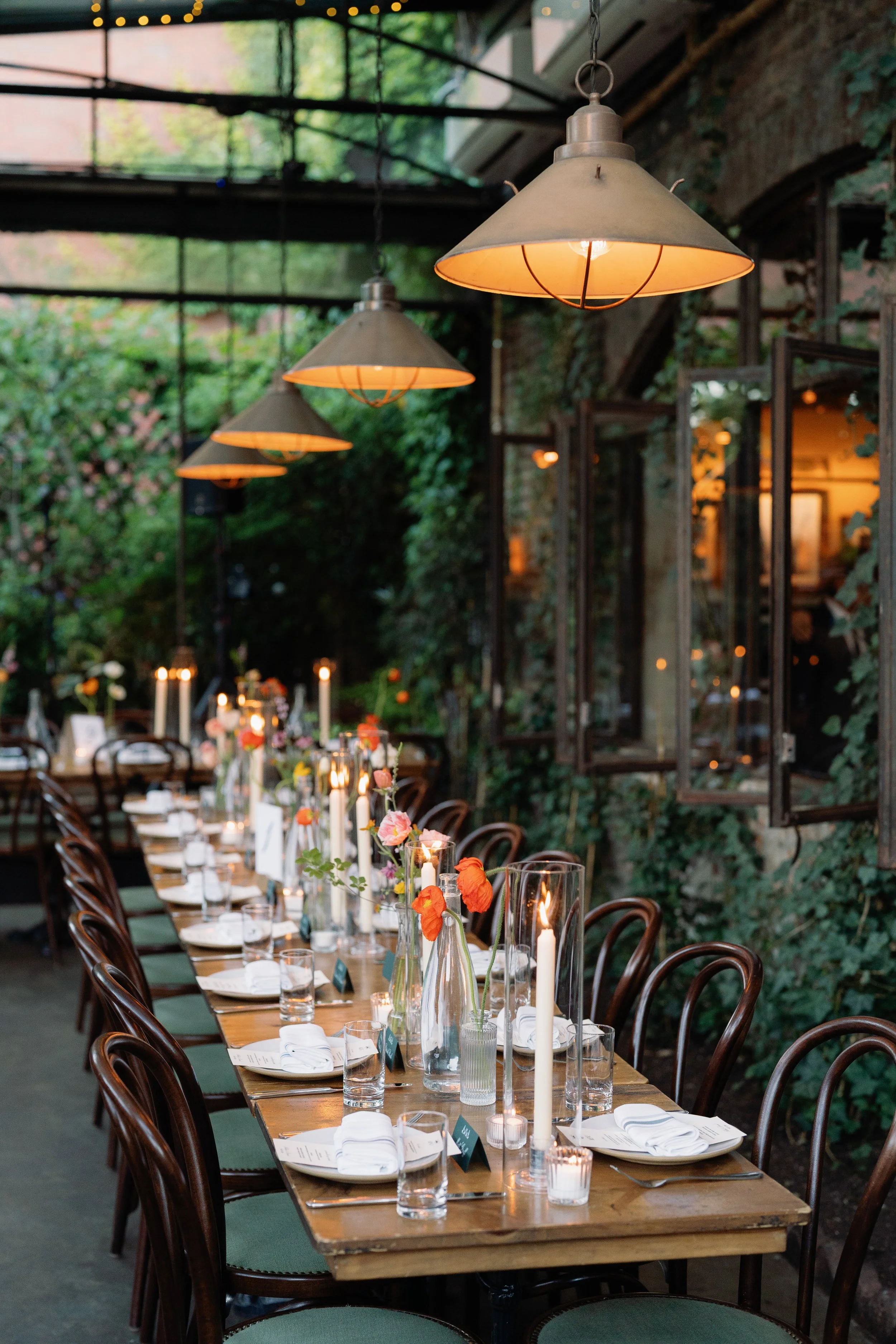 A long wooden dining table set for a formal event with candles, flowers, and glasses in a greenhouse-like space with hanging lamps and greenery outside.