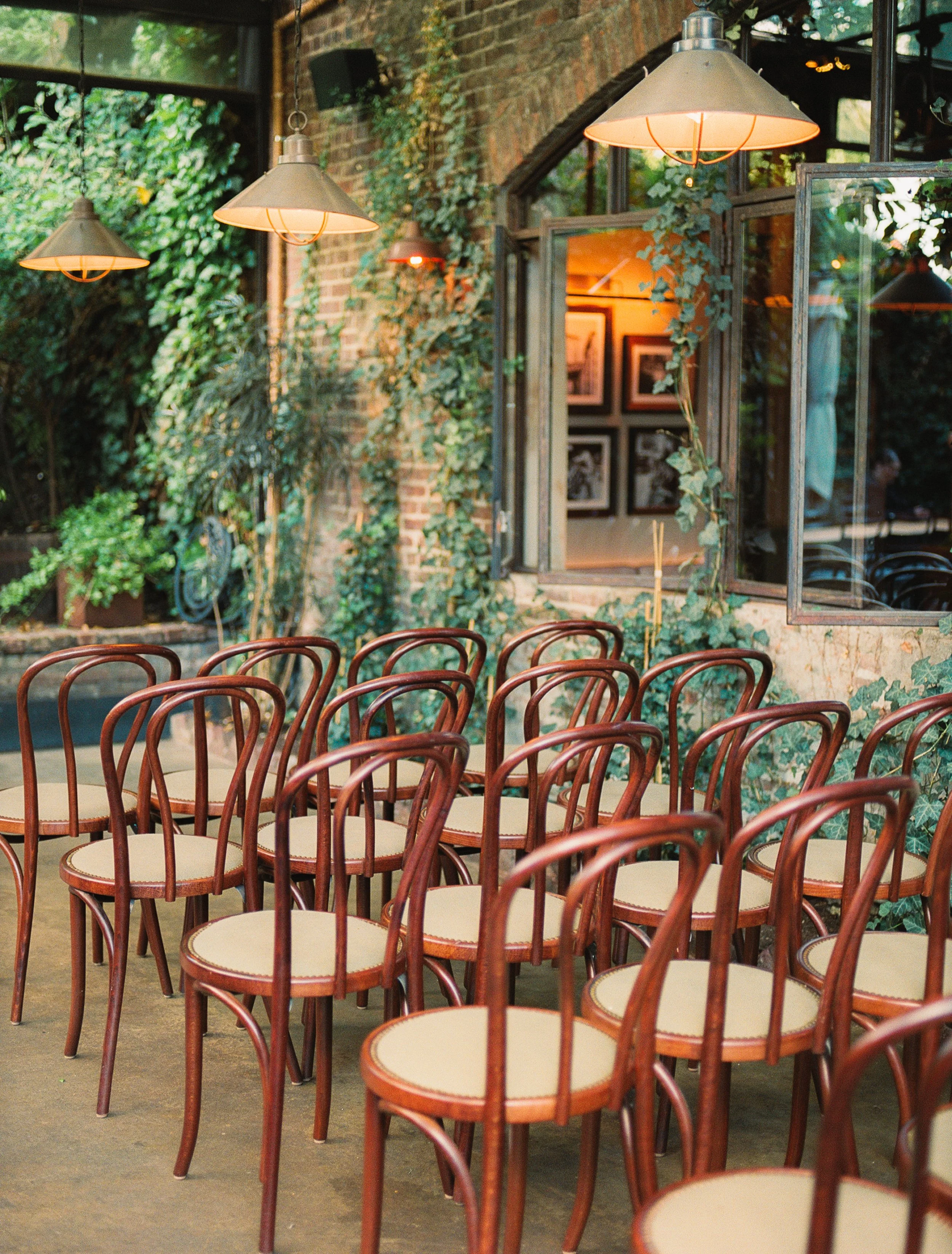 Empty wooden chairs arranged around a table inside a rustic indoor space with brick walls, hanging lights, and green plants.