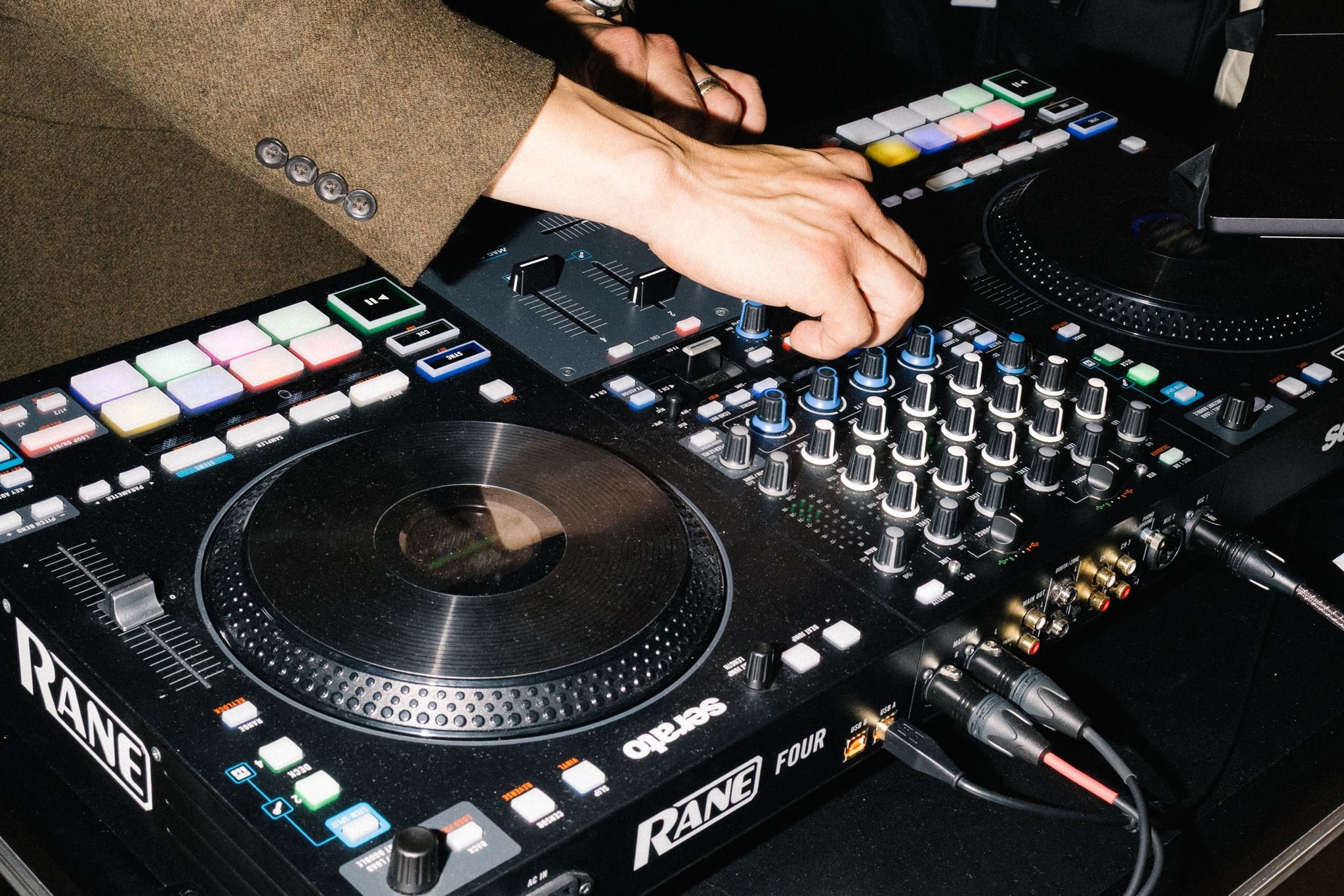 Close-up of a DJ's hands operating a professional DJ mixer and turntable, with colorful buttons and knobs, in a dark setting.