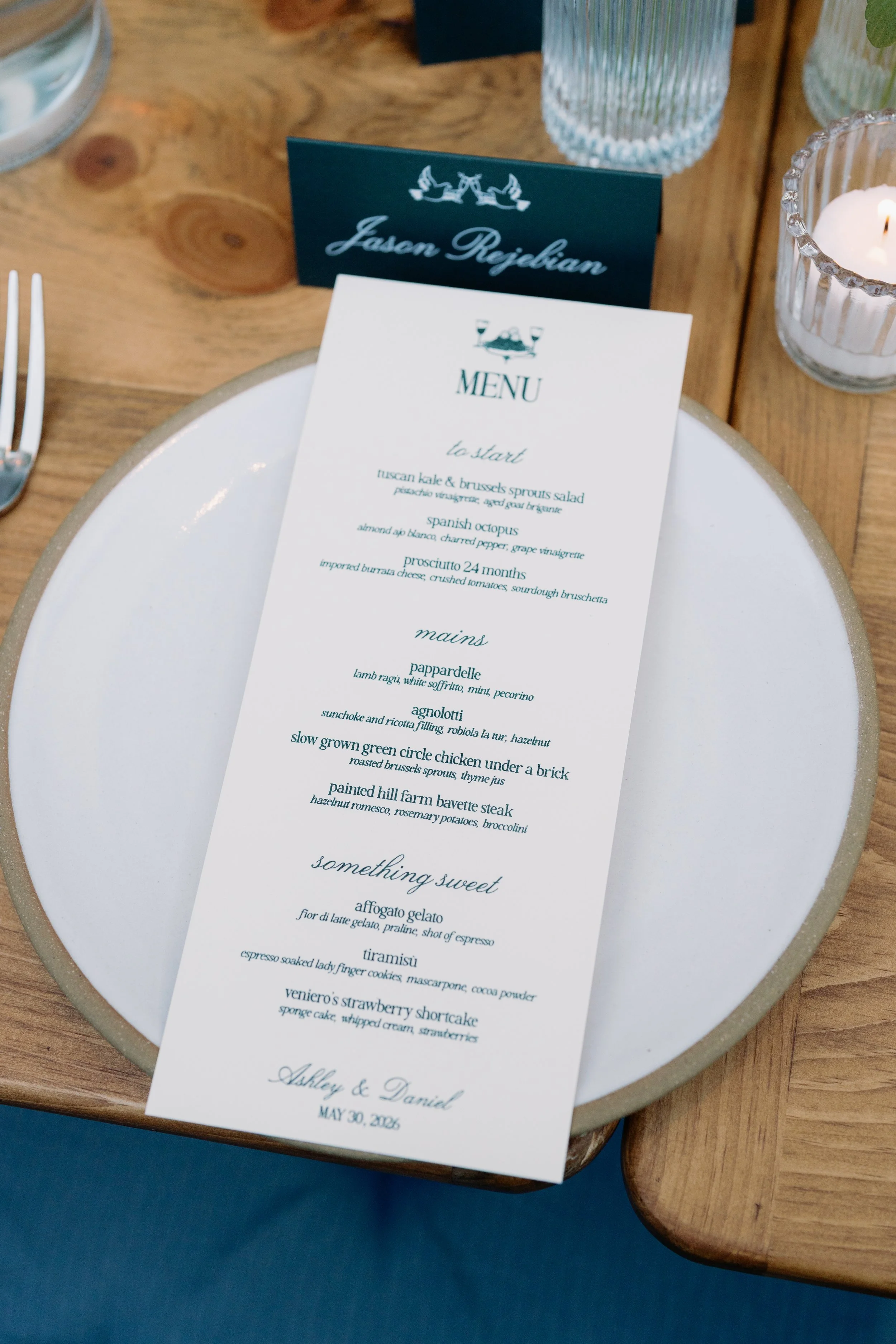 A wedding menu reading 'Jason Riedyian' and 'Ashley & Daniel' with the date May 30, 2023, on a table with a white plate, a glass of water, a lit candle in a glass holder, and a place card.