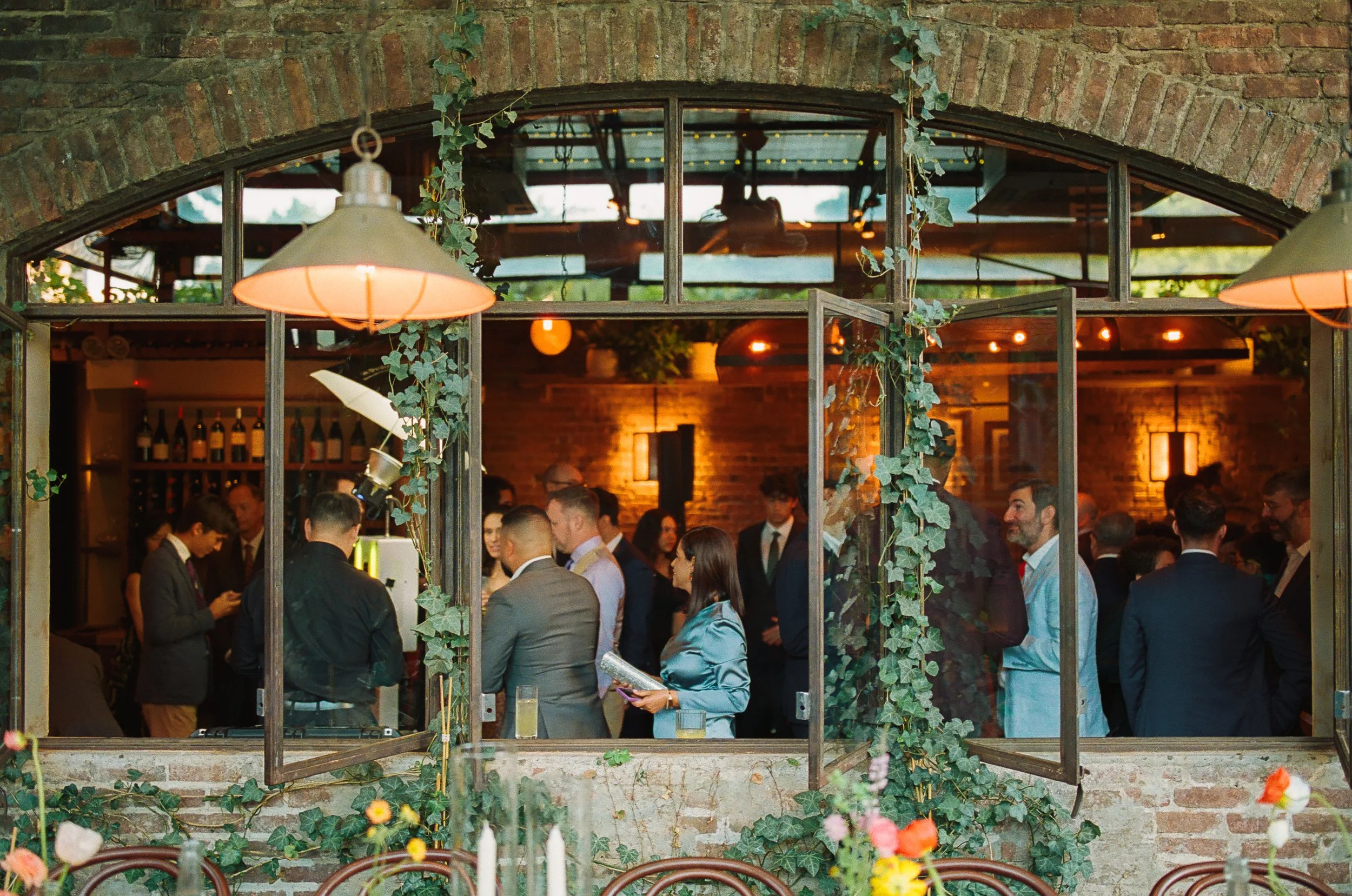 People socializing inside a rustic, brick-walled restaurant or event space, viewed through a window adorned with ivy and plants, with warm lighting and a bar area in the background.