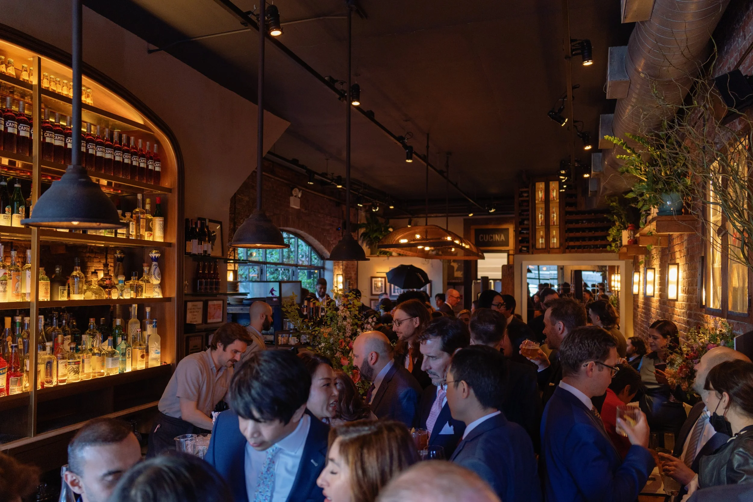 A crowded bar or restaurant filled with people socializing. Shelves of liquor bottles and hanging pendant lights are visible, with exposed brick walls and large windows in the background.