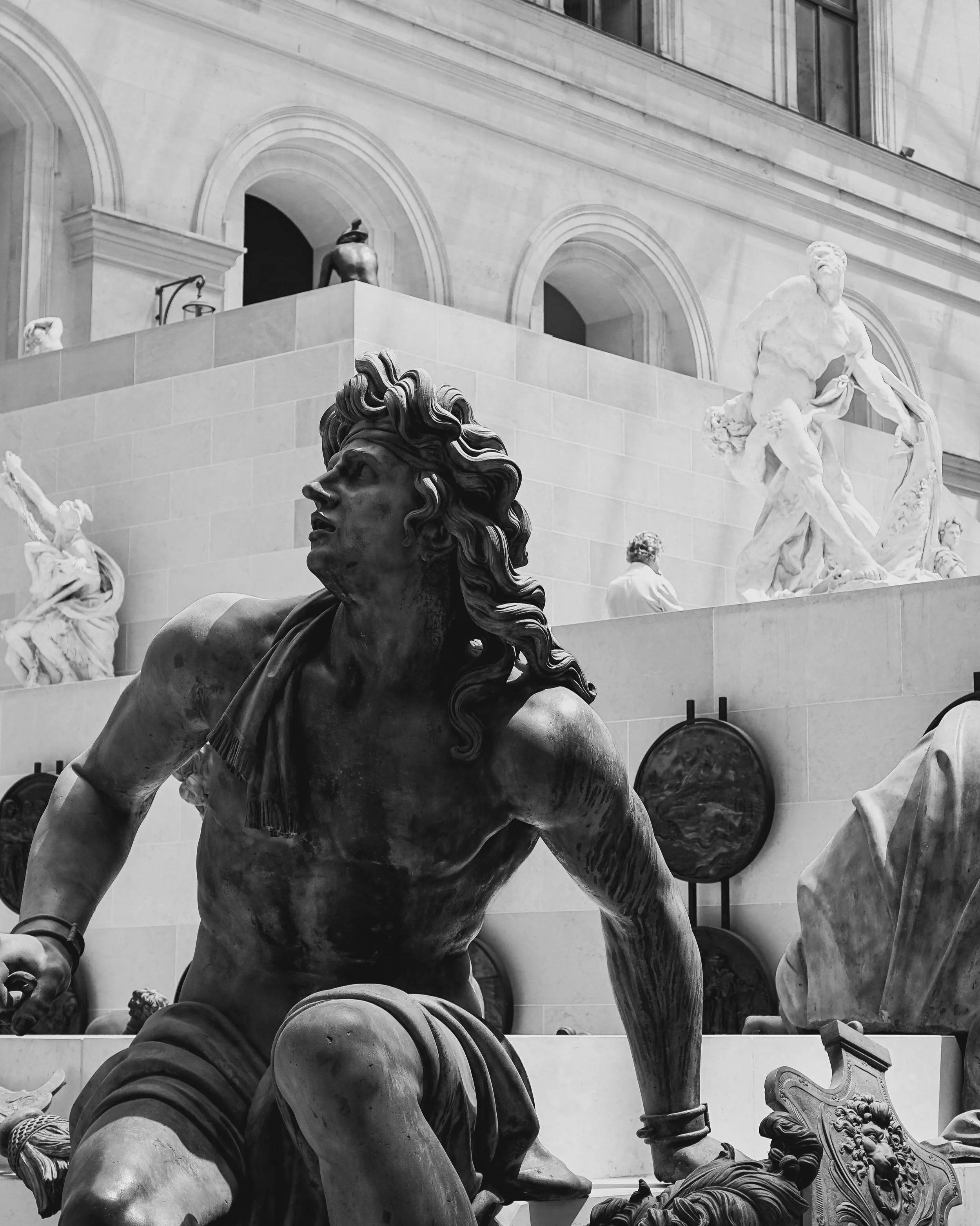Black and white photo of a classical statue depicting a muscular figure in motion, surrounded by other sculptures and decorative elements in a museum setting.