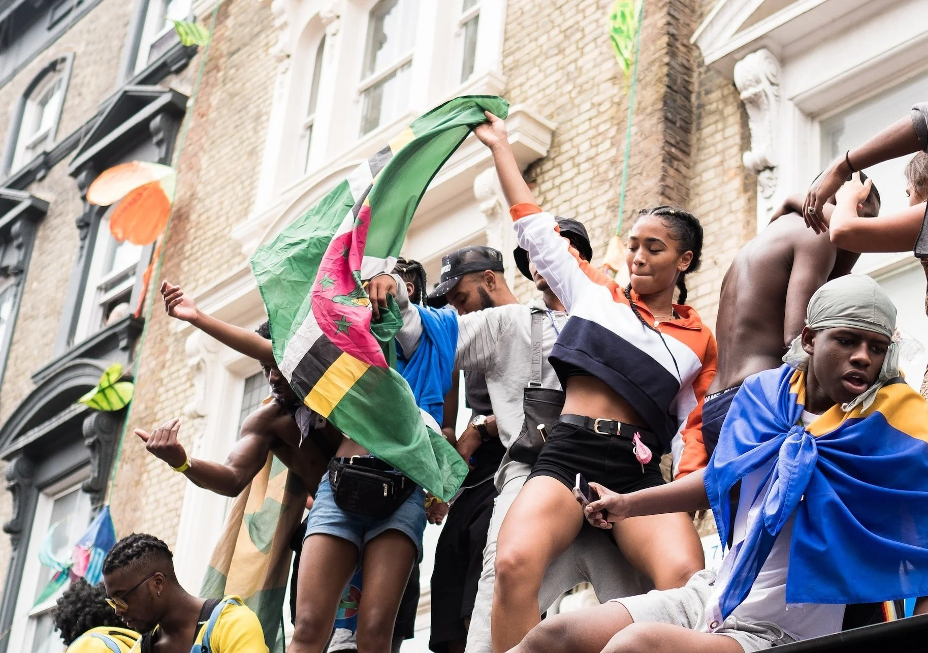 People dancing and holding flags in a street festival, colorful buildings in the background.