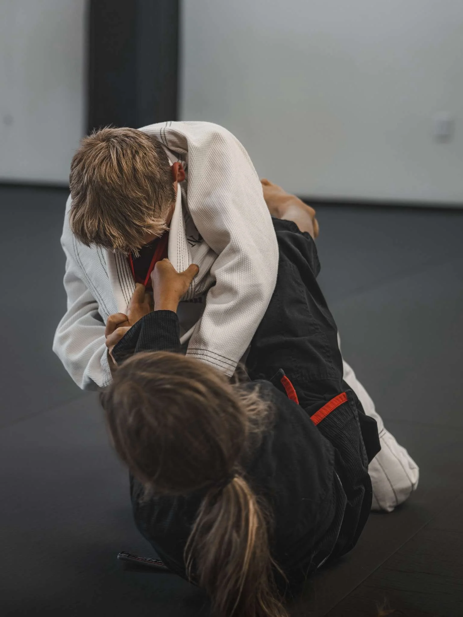 Two kids practicing Brazilian Jiu-Jitsu on a black mat, wearing gi uniforms, in a martial arts gym.