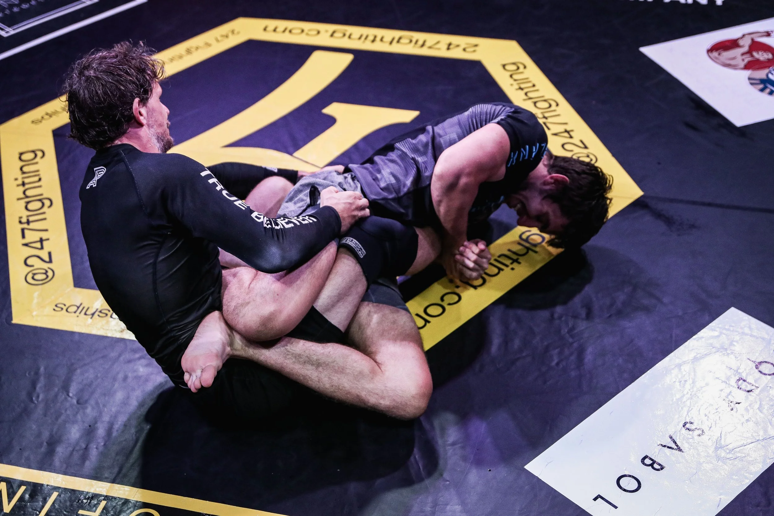 Two men are grappling on a black and yellow mixed martial arts mat. One man is on his back in a defensive position, while the other is on top, attempting control. The mat features a large yellow logo and text surrounding the area.