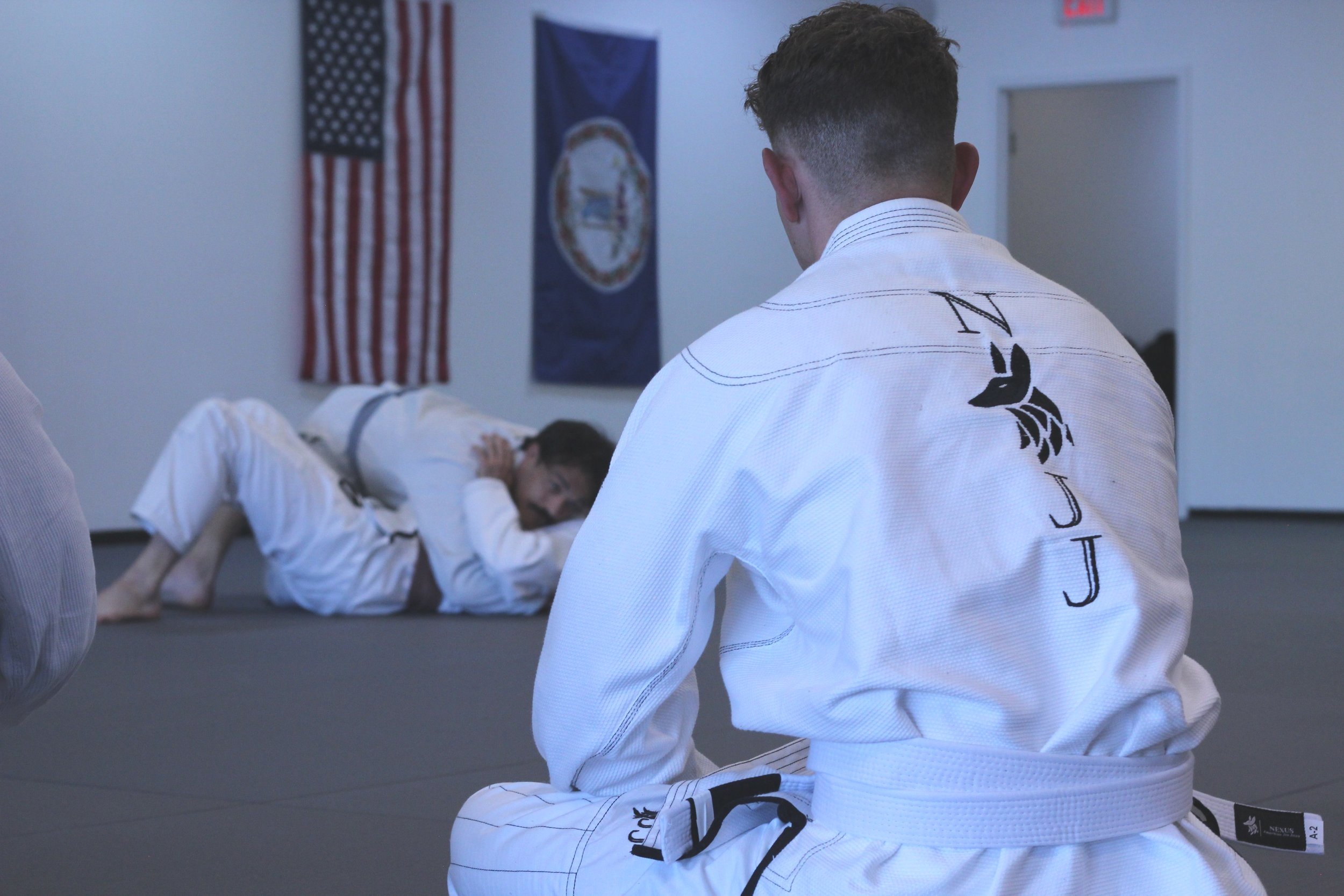 A person in a white gi sitting cross-legged on a martial arts mat, observing a class. Beginner course for jiu jitsu in Fredericksburg Va.