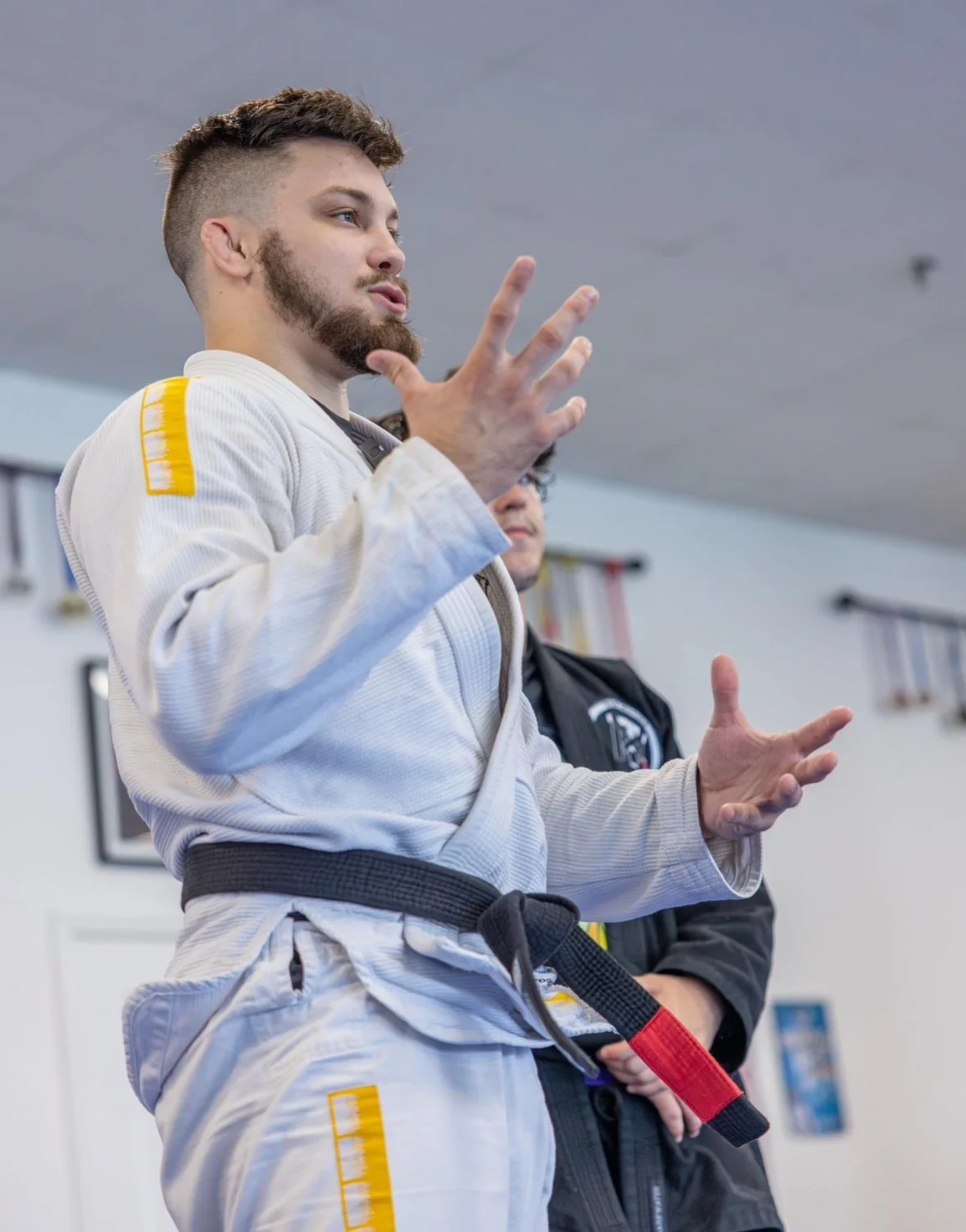 A man in a white Brazilian Jiu-Jitsu gi with a black belt, speaking and gesturing with his hands in a martial arts training facility.