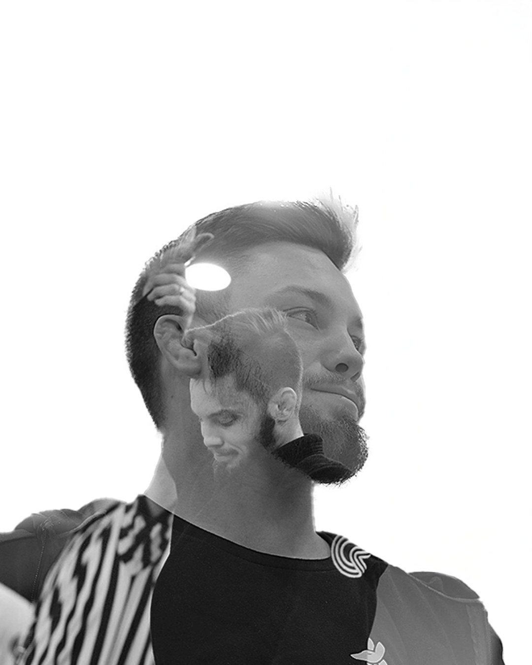 Black and white double exposure photo of a man with short hair and a beard, smiling slightly, wearing a dark shirt, with reflections of himself and a woman in a striped shirt visible on his face and shoulder.