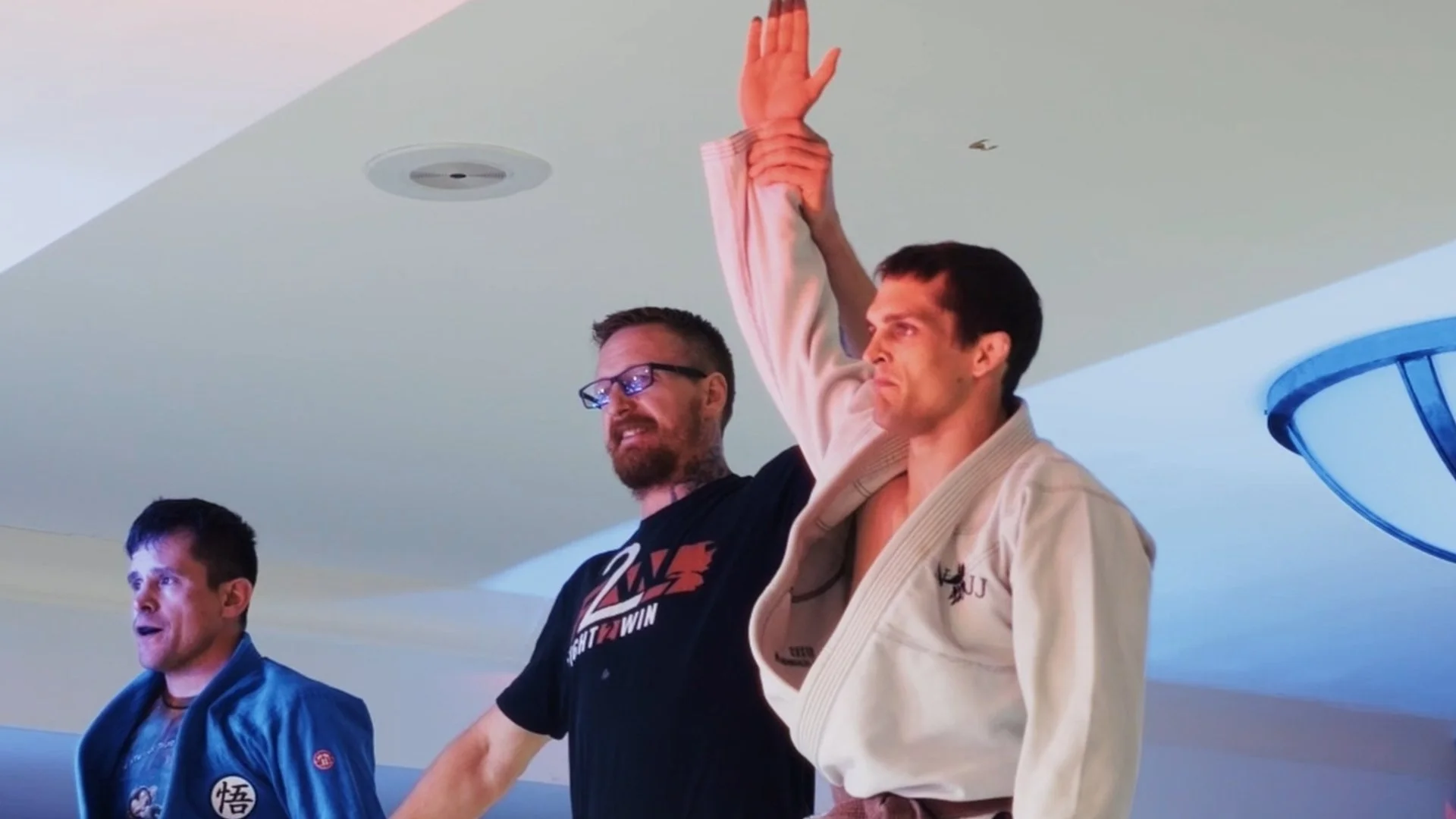 Three men participating in a Brazilian Jiu-Jitsu seminar, one in traditional gi, one in casual clothing, with a hand raised in the air.