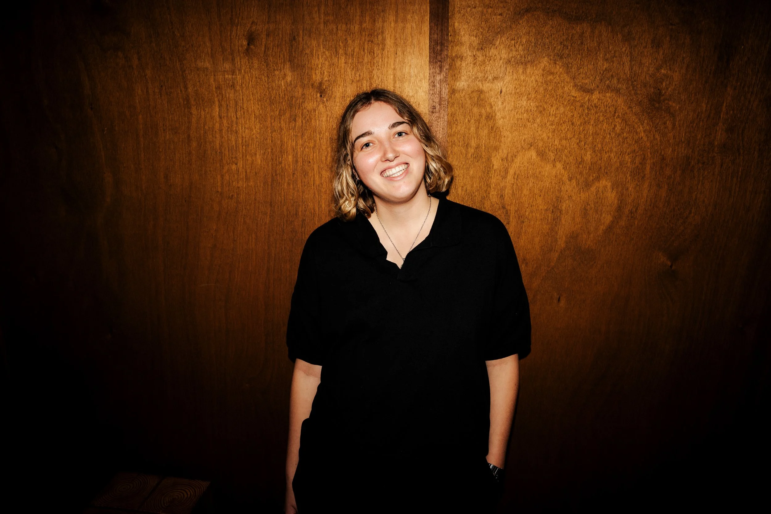 A young woman with shoulder-length blonde hair and a black shirt stands against a wooden wall, smiling at the camera.