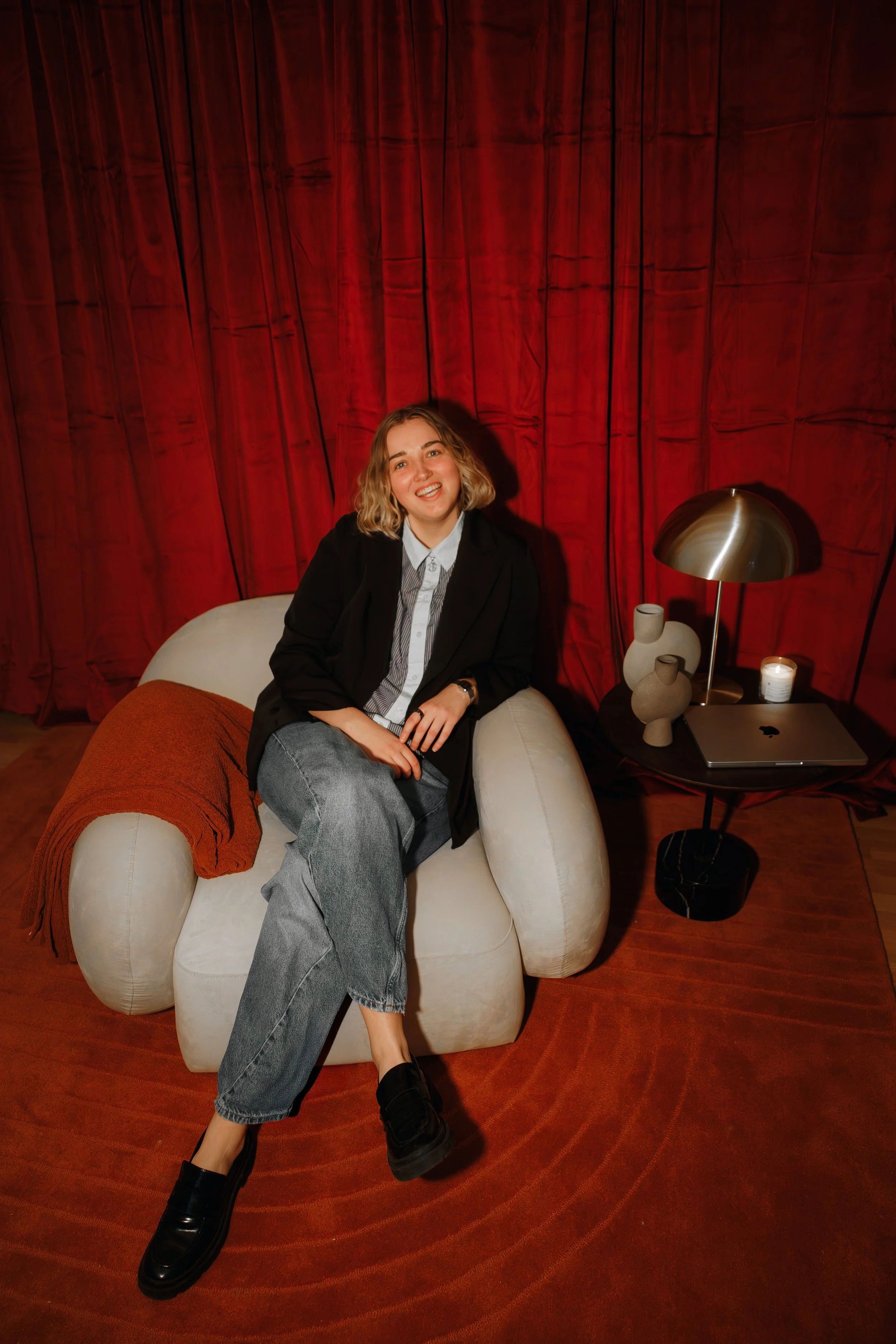 A young woman with blonde hair, smiling, sitting on a white, rounded armchair with a rust-colored blanket draped over it. She is wearing a black blazer, a striped shirt, and gray jeans. Behind her is a red curtain. To her right, there is a small black table with a metallic lamp, three vases, a lit candle, and a closed laptop. The floor is wooden, and the overall setting appears cozy and stylish.