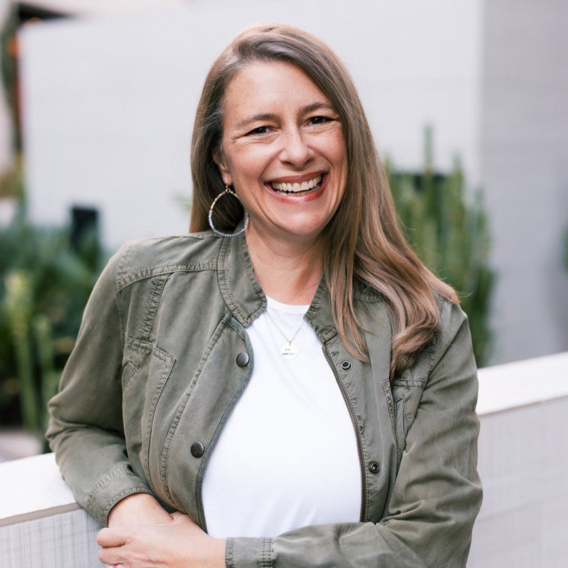 Smiling woman with long brown hair wearing hoop earrings, a white t-shirt, and an olive green jacket, standing outdoors with a blurred background of plants and a building.