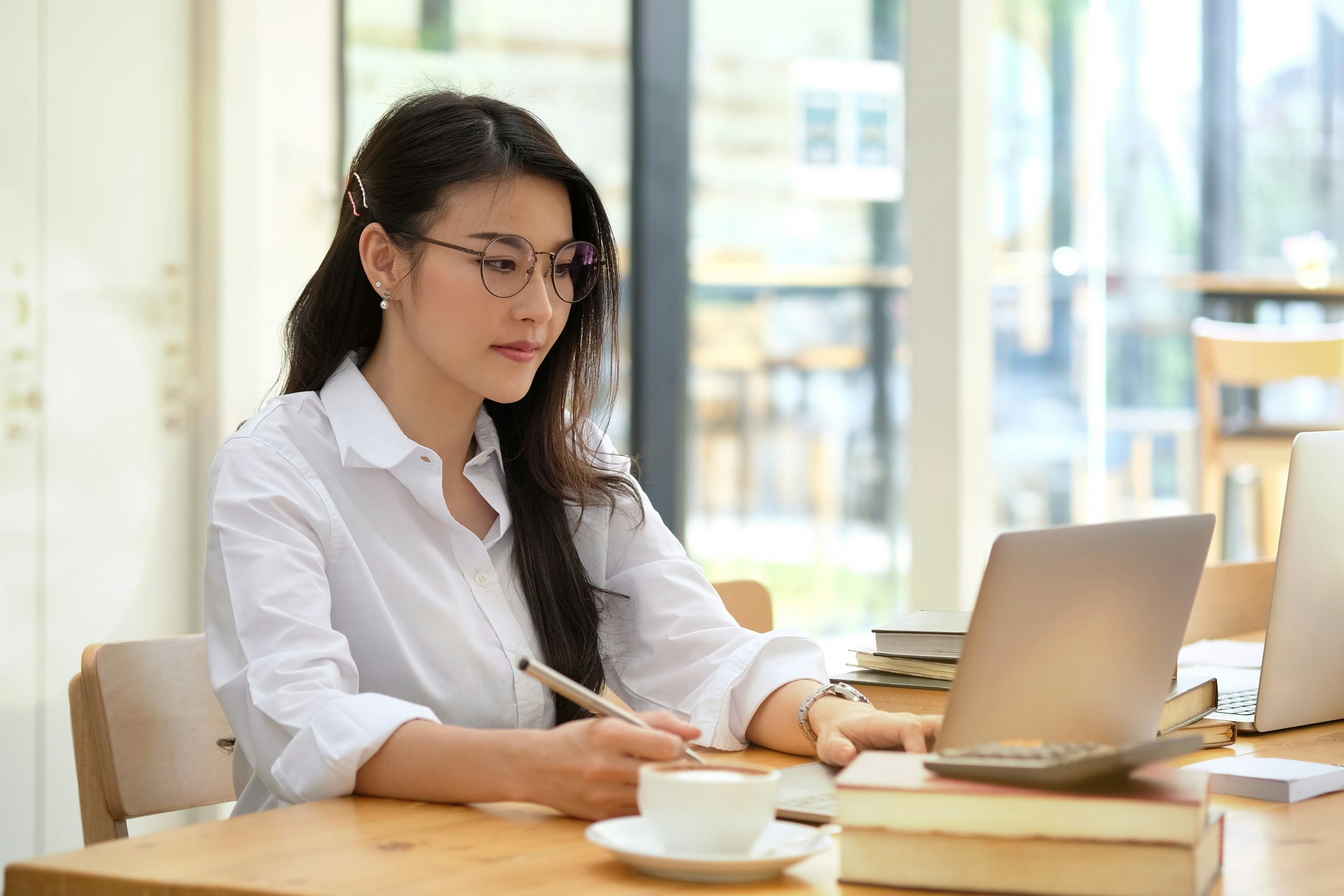 Woman with long dark hair, glasses, and pearl earrings working at a wooden table with laptops, books, and a cup, sitting in a bright modern café or workspace.