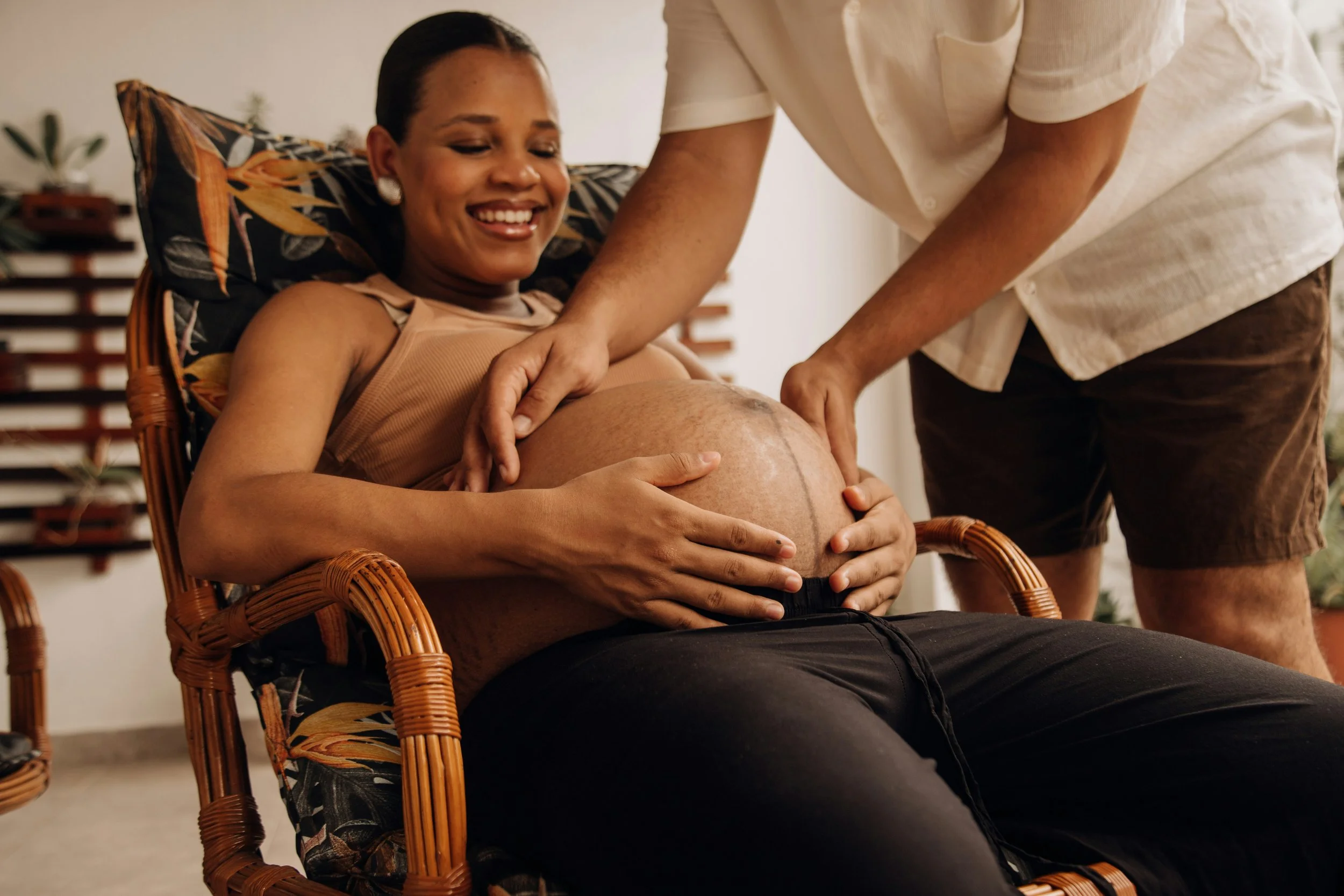 A pregnant woman sitting in a chair with a floral cushion, smiling as she touches her baby bump while a man, partially visible, gently places his hands on her belly.