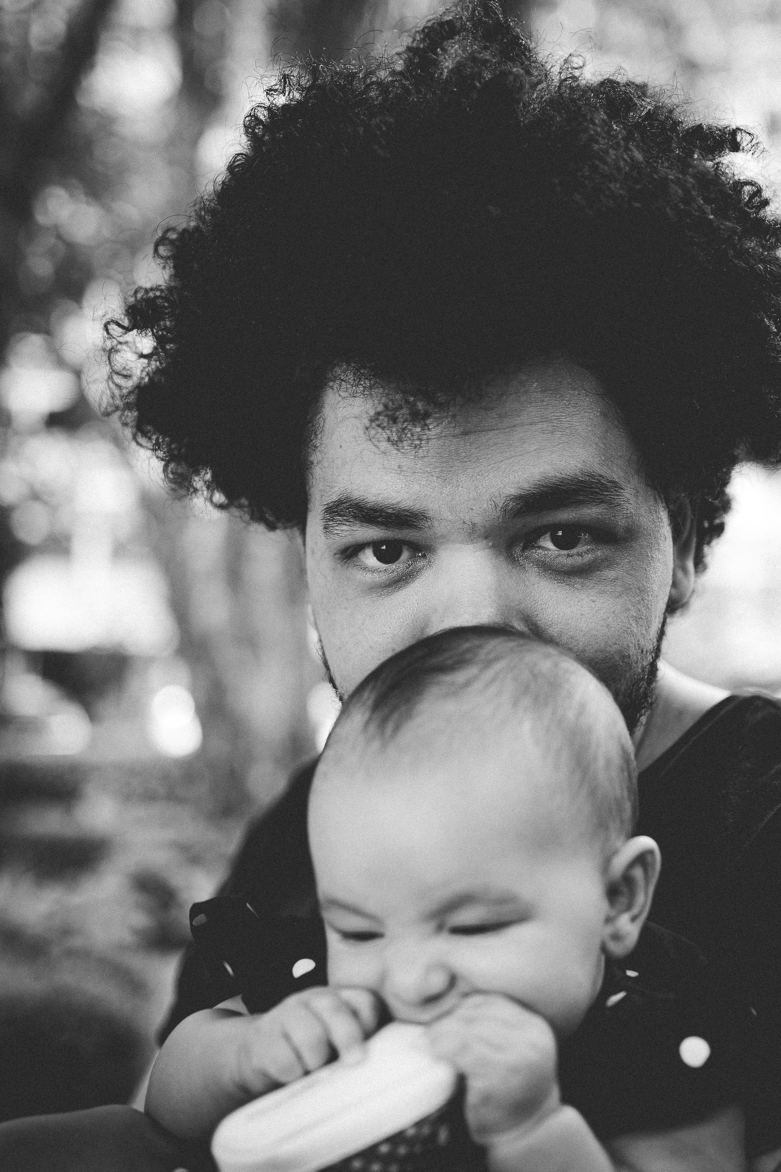 A black-and-white close-up photo of a man with curly hair and a beard holding a baby, with the baby chewing on a toy or object.