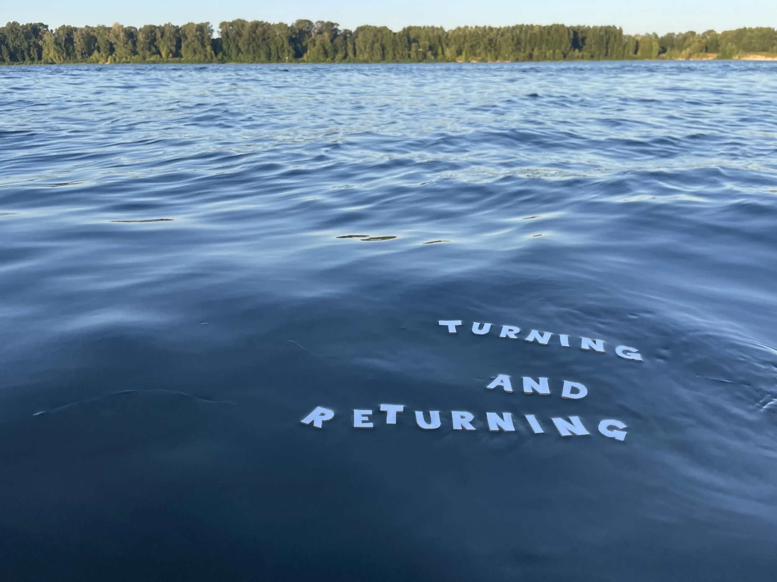 A body of water with gentle waves, a distant tree-lined shoreline, and partially visible white text on the water that reads 'TURNING AND RETURNING'.