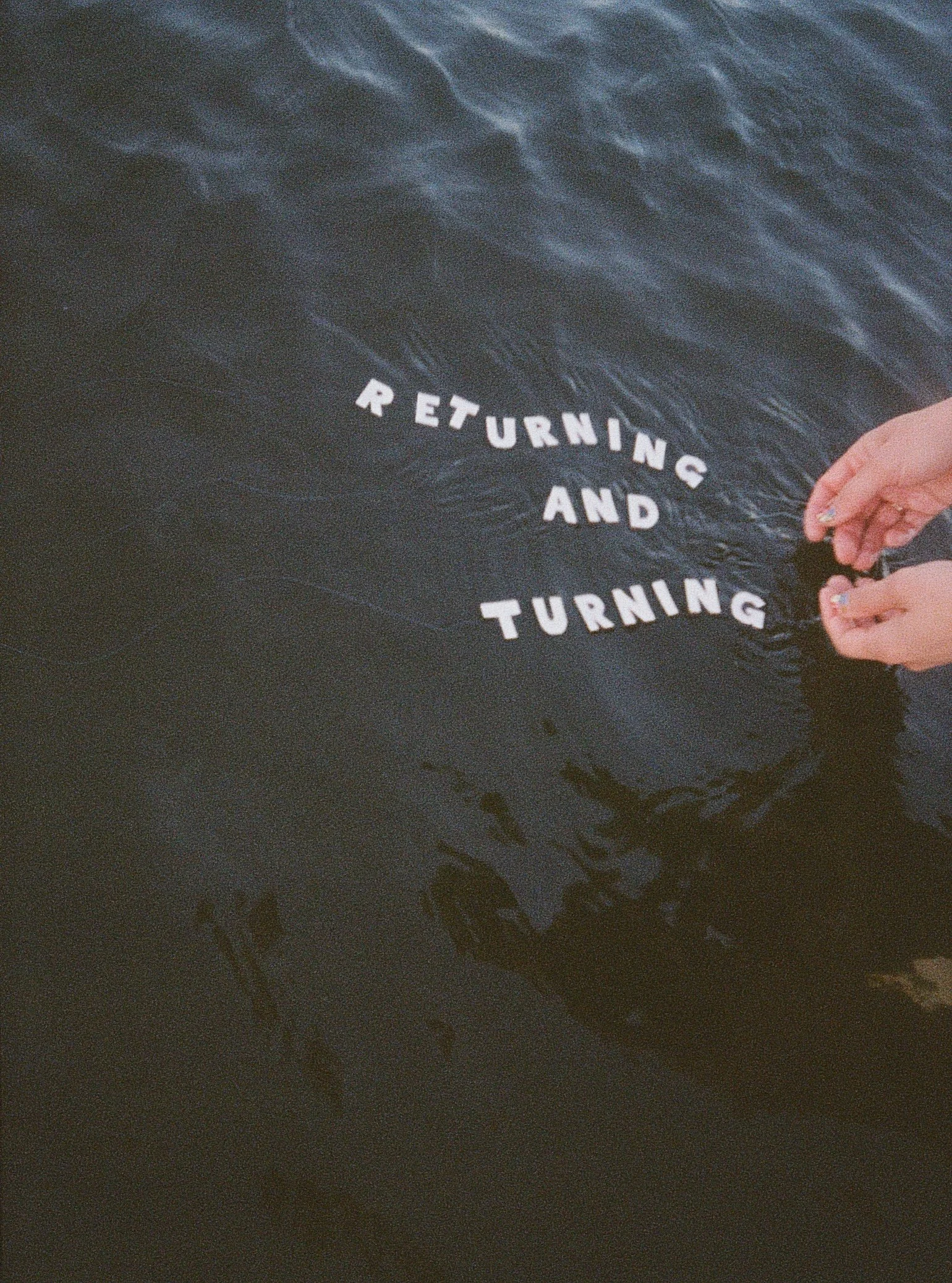 A person's hand holding white letter cutouts spelling out "RETURNING AND TURNING" against a dark water background.