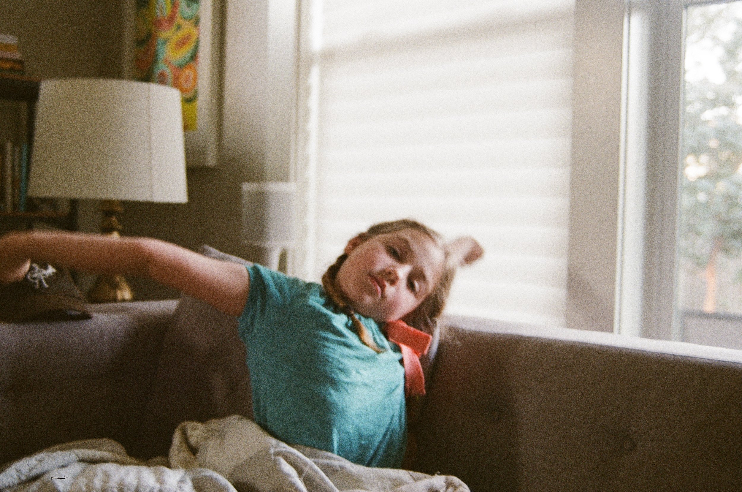 A young girl with braided hair, wearing a teal shirt, is relaxing on a brown couch near a window with white blinds, in a cozy living room with a bookshelf and a lamp in the background.