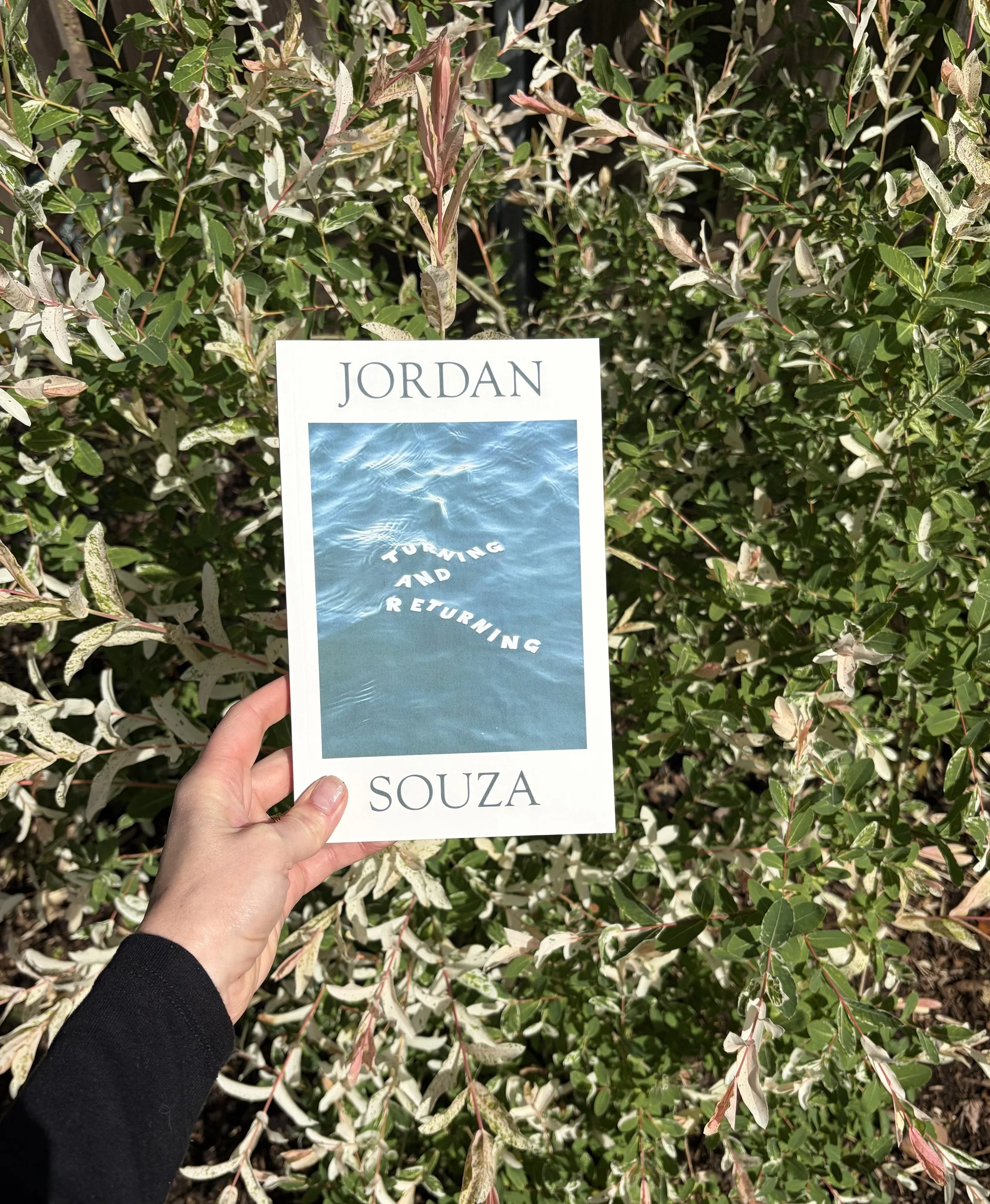 Hand holding a paper sign in front of a lush leafy bush. The sign shows the word "JORDAN" and the name "SOUZA" with an image of water and a path of stepping stones.