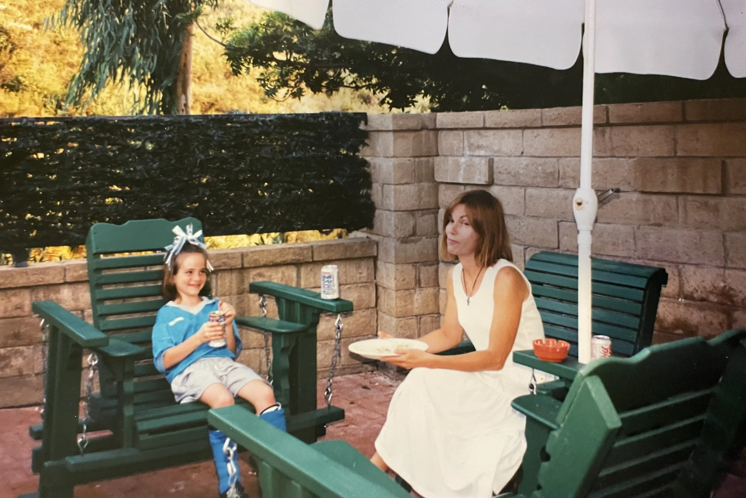 A woman and a young girl sitting on green outdoor chairs at a table on a patio, surrounded by brick walls and a white patio umbrella. The girl is smiling and holding a drink, while the woman is looking at the camera and holding a plate.