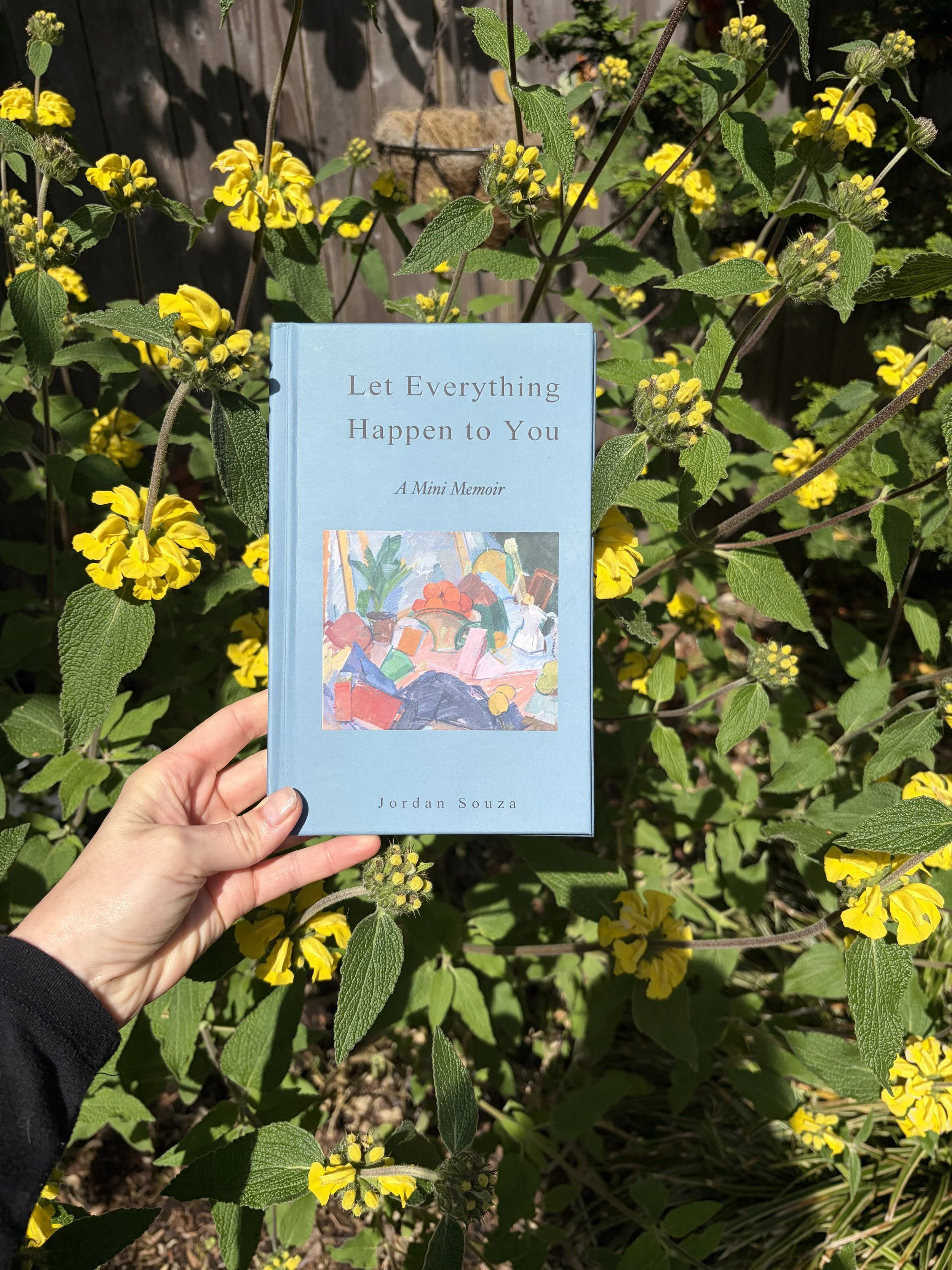Hand holding a light blue book titled "Let Everything Happen to You: A Mini Memoir" by Jordan Souza in a garden with yellow flowers and green leaves, with a wooden fence in the background.