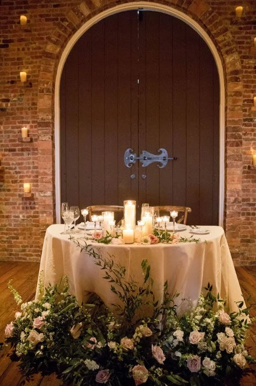 Elegant wedding table setting with candles and floral arrangements against a brick wall and arched wooden door.