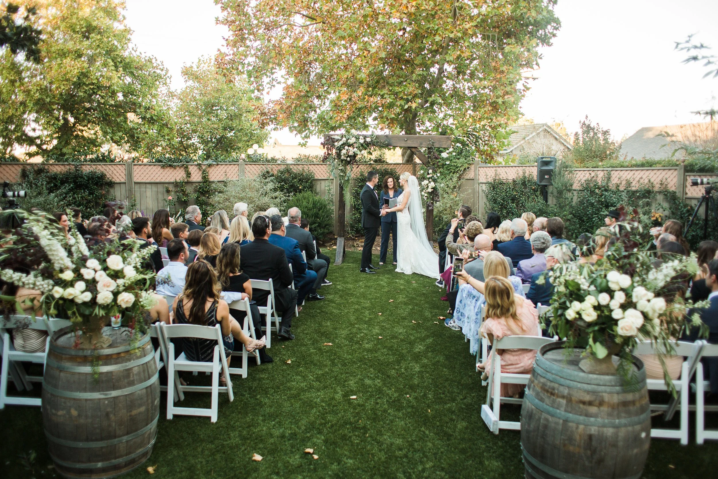 Outdoor wedding ceremony with couple under wooden arch, officiant, guests seated, surrounded by greenery and decorative floral arrangements on barrels.