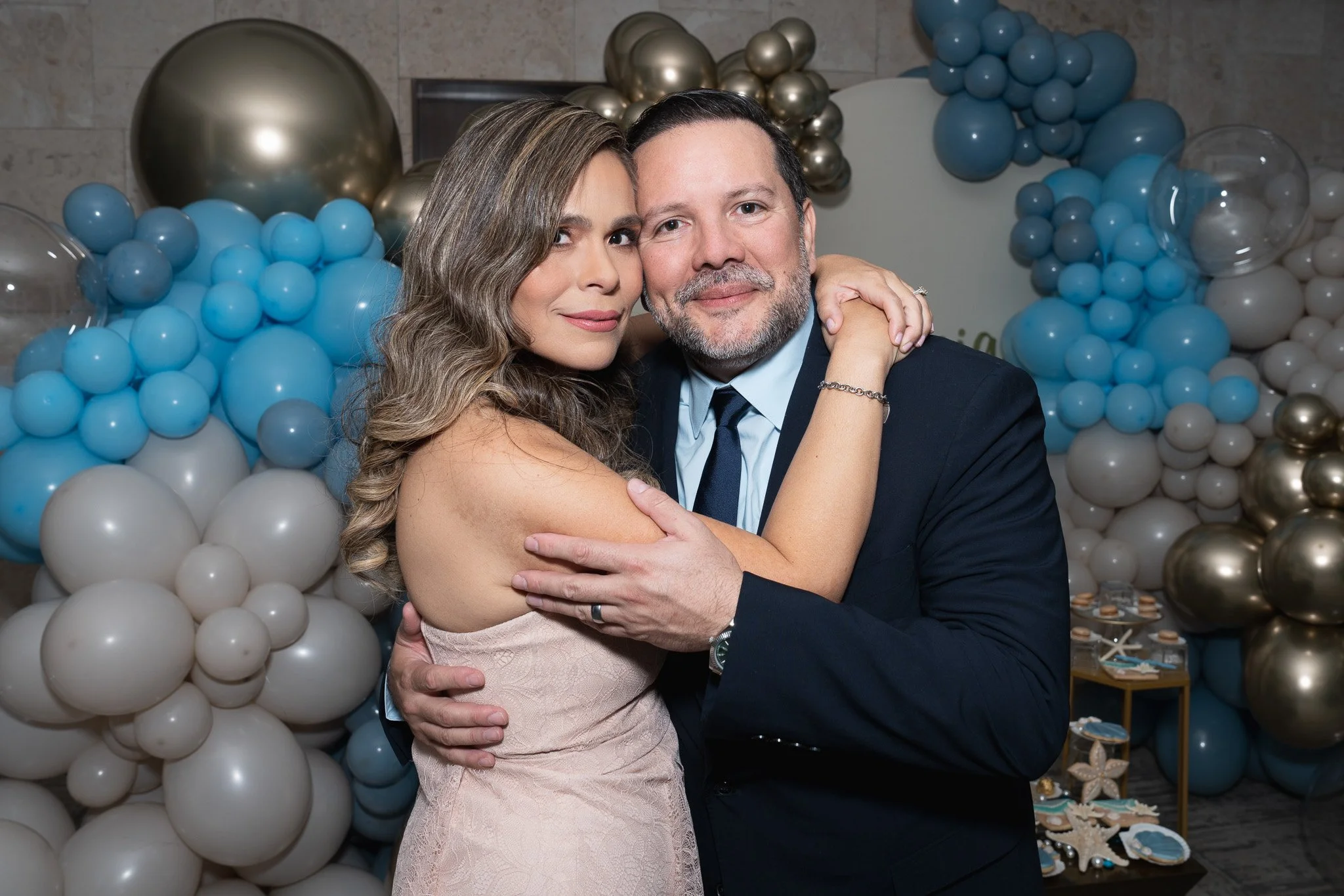 A woman and a man are embracing each other and posing for a photo at what appears to be a celebration, with a backdrop of blue, white, and gold balloons.