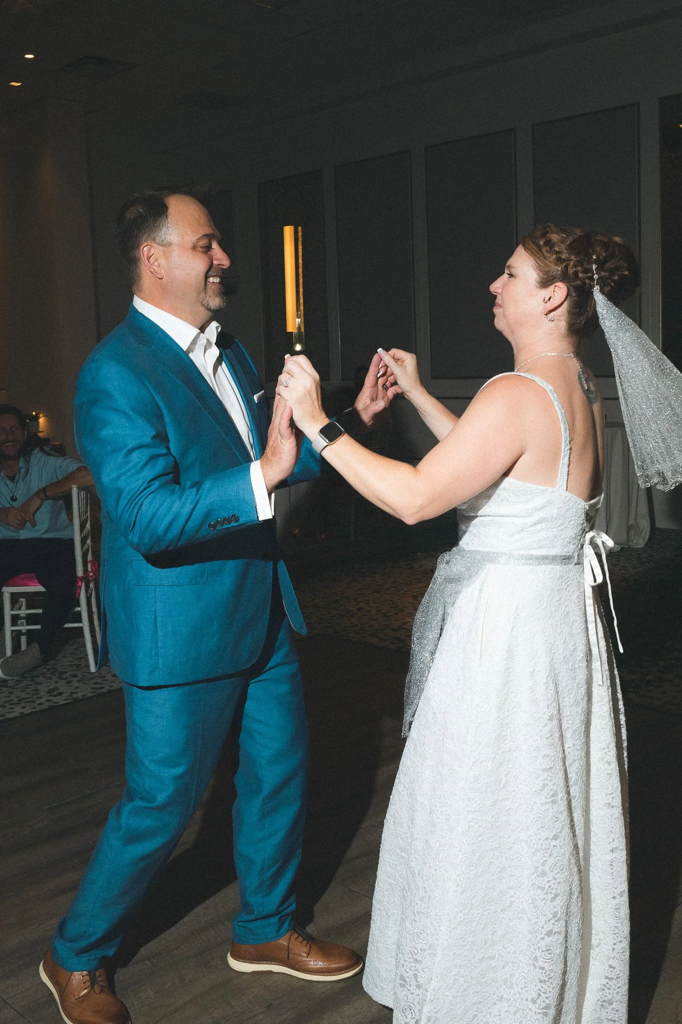 A bride and groom exchanging wedding rings during their ceremony. The groom is wearing a blue suit and the bride is in a white wedding gown with a veil. They are smiling and holding hands, in an indoor reception area.