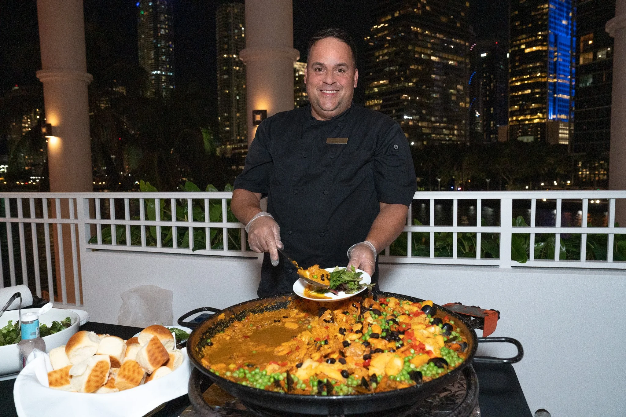 A chef serving a large pot of colorful paella at an outdoor event at night with city skyscrapers in the background.