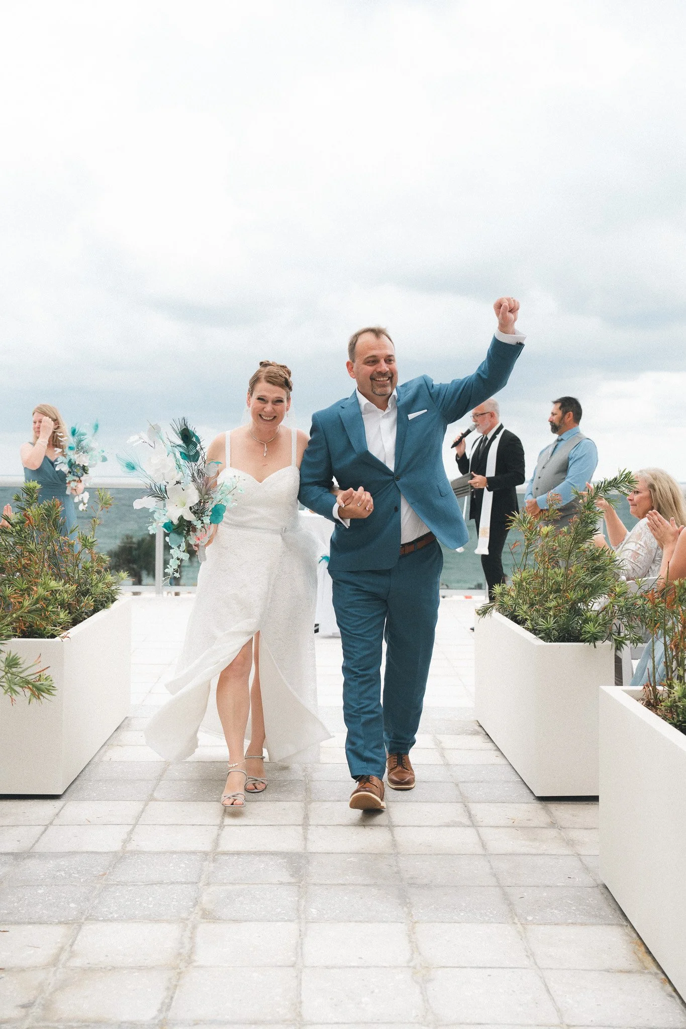 A newly married couple walking down an outdoor aisle, smiling and celebrating, with friends and family clapping around them, on a cloudy day.