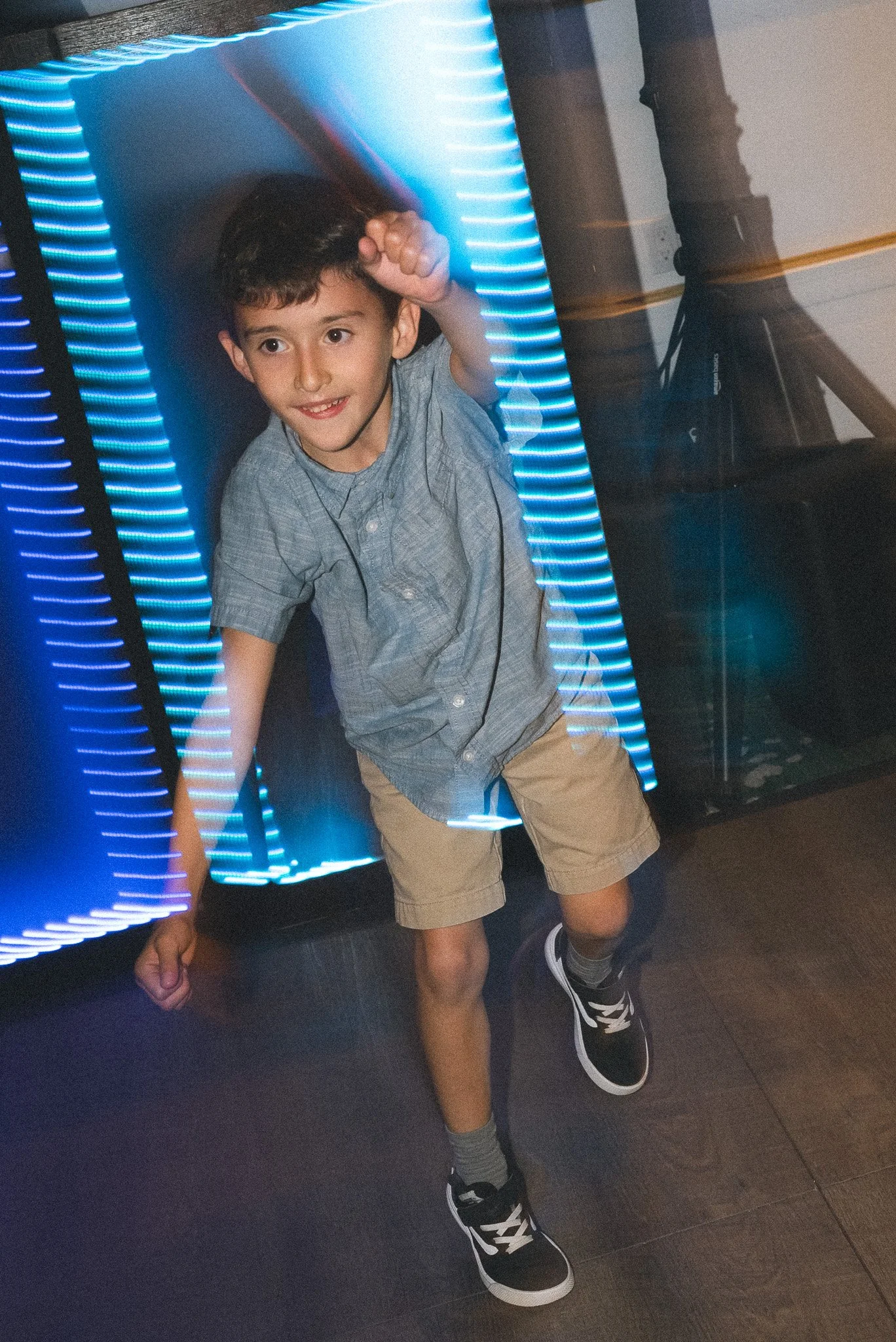 A young boy with curly brown hair and a gray shirt posing playfully inside a tunnel with blue neon lights at an arcade or entertainment venue.