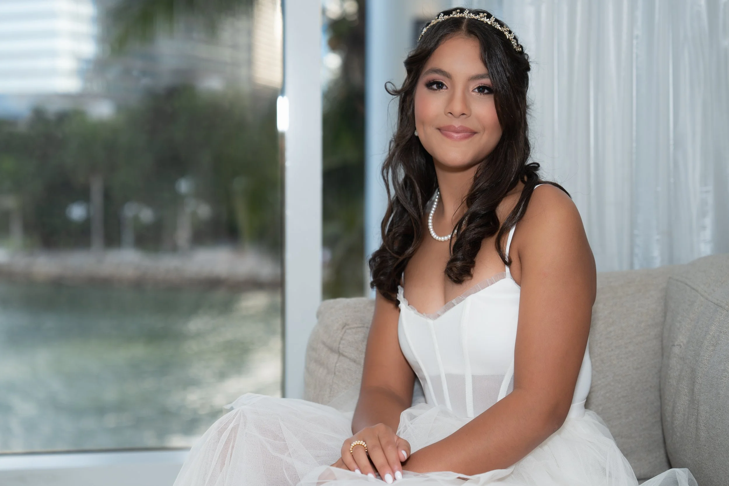 Young woman in a white dress with pearl jewelry and a headband sitting on a couch near a window looking at the camera.