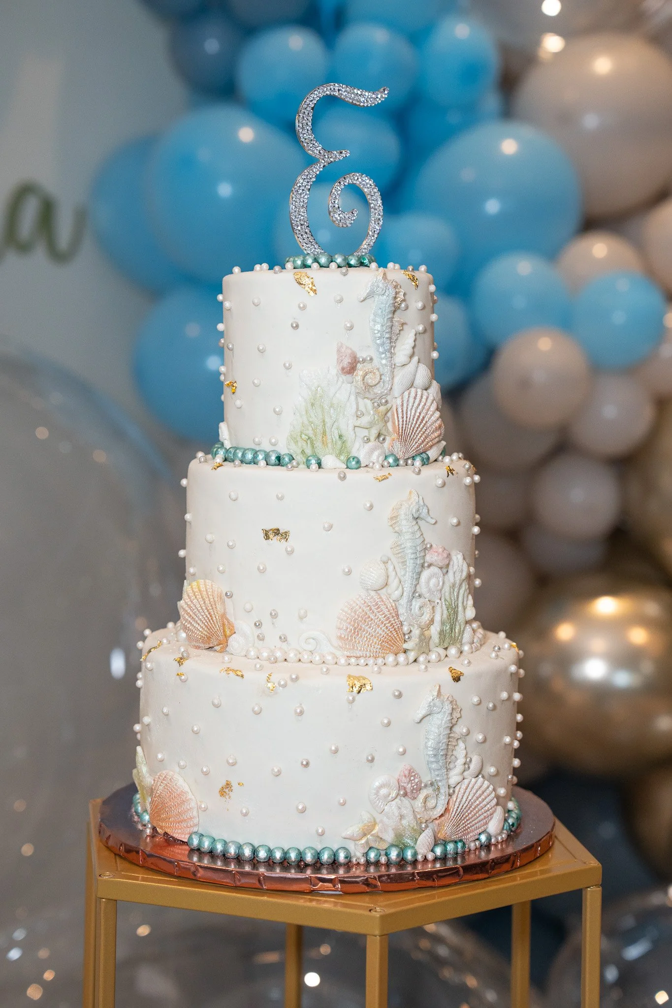 A three-tiered wedding cake decorated with seahorses, seashells, and pearls, topped with a sparkling silver hook shaped cake topper, placed on a gold stand with a backdrop of blue and white balloons.