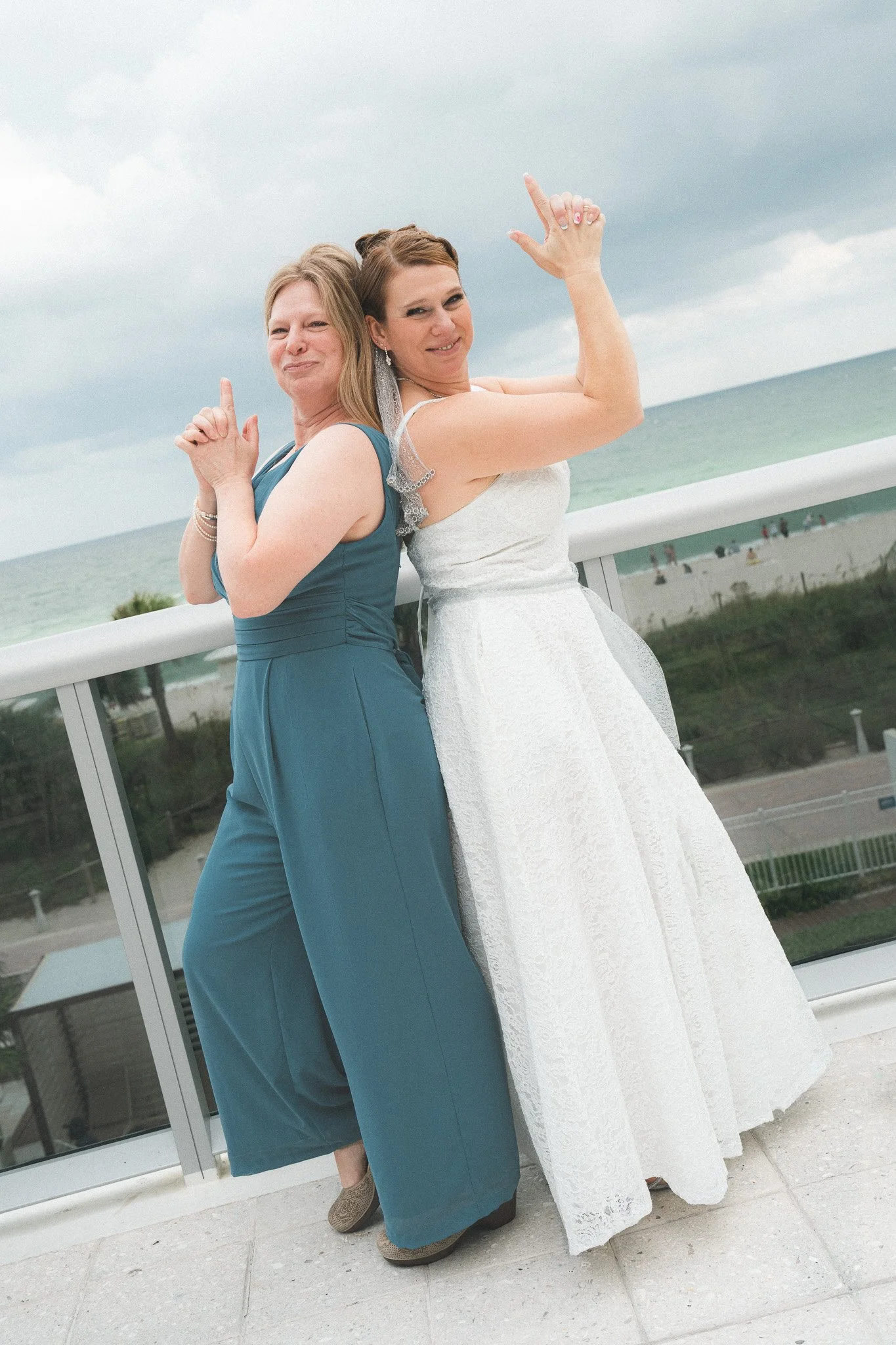 Two women, one in a white wedding dress and the other in a teal dress, standing back to back on a balcony overlooking a beach, posing playfully.