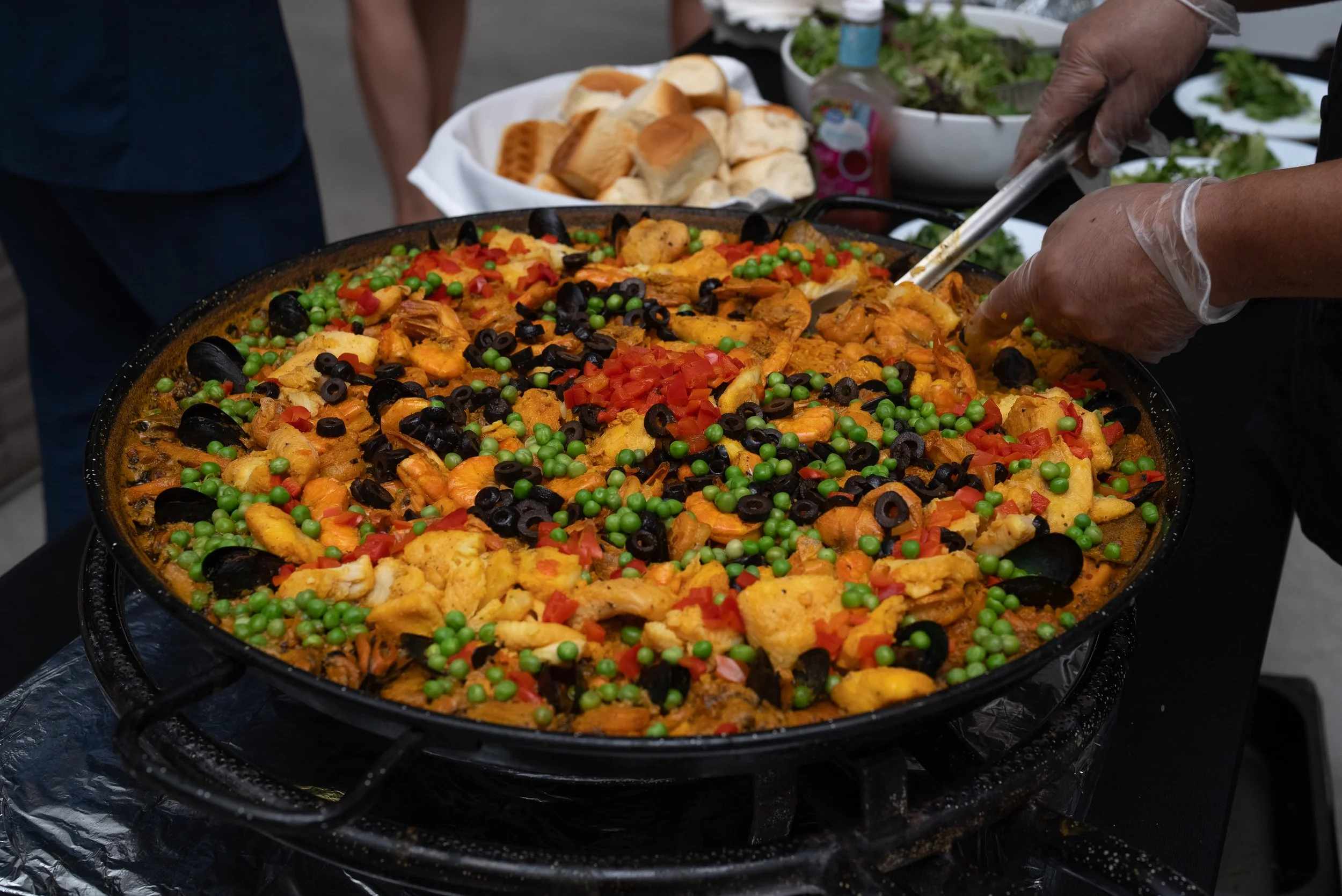A large pan of seafood paella with shrimp, mussels, peas, red peppers, and rice, being served at an outdoor event with bread and salads on the side.